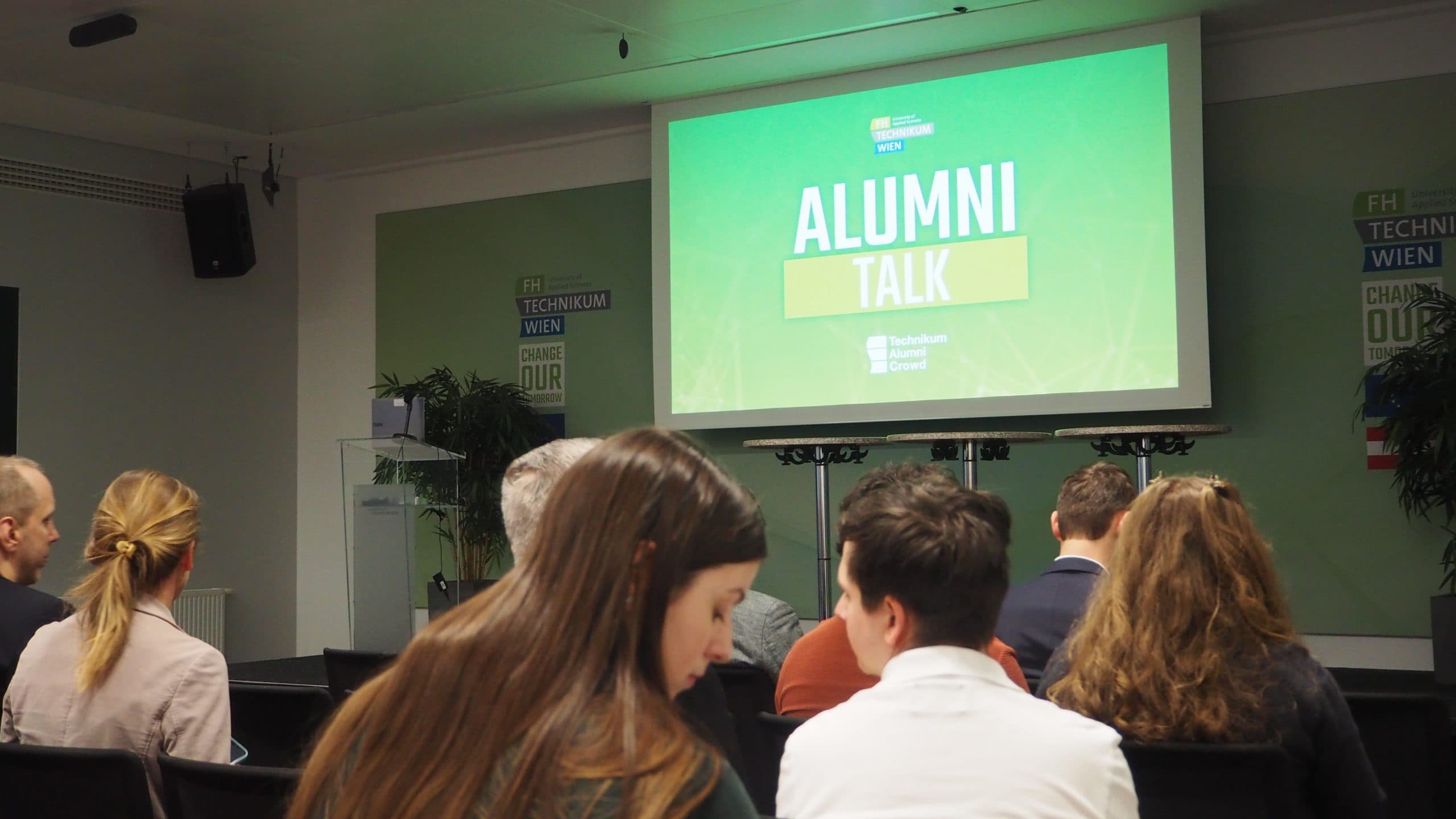 Die Zuhörer sitzen vor einem großen Bildschirm mit der Aufschrift 'ALUMNI TALK' in einem Konferenzraum. Die Atmosphäre ist ruhig, während die Menschen auf den Beginn der Veranstaltung warten.