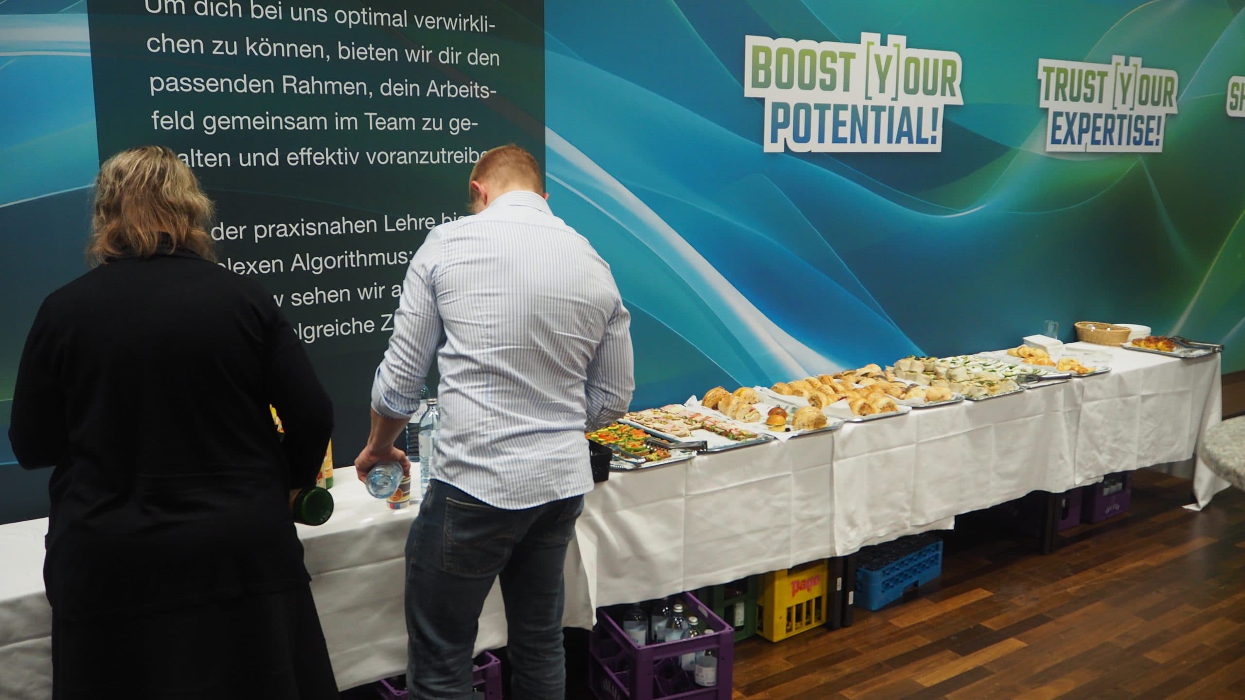 Zwei Personen stehen an einem langen Tisch mit verschiedenen Frühstücksspeisen in einer Konferenzumgebung. Hinter ihnen sind blaue und grüne Banner mit den Aufschriften BOOST YOUR POTENTIAL! und TRUST YOUR EXPERTISE! zu sehen.