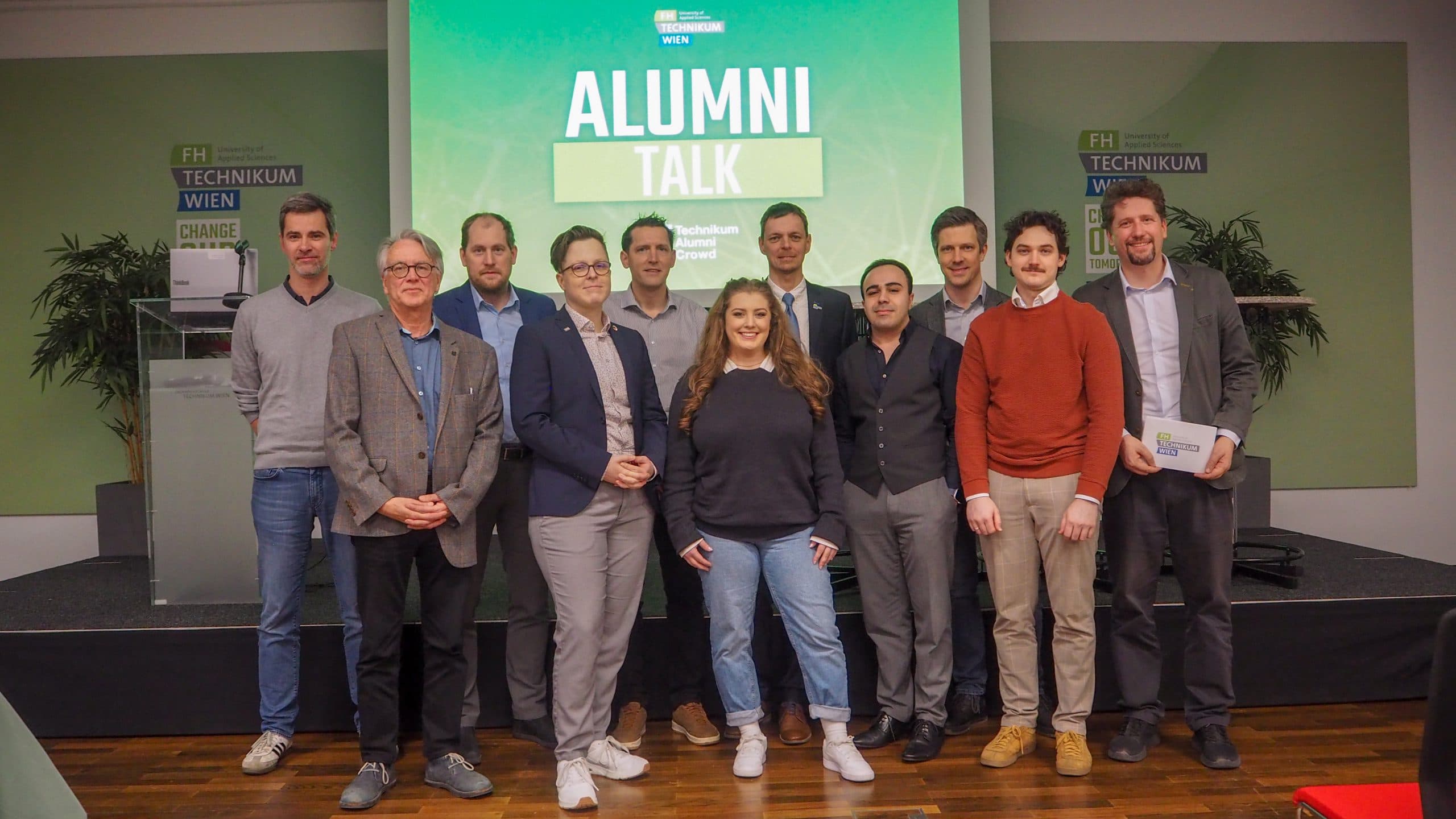 Eine Gruppe von zwölf Personen steht Seite an Seite auf einer Bühne und lächelt in die Kamera. Hinter ihnen ist ein Greenscreen mit der Aufschrift 'ALUMNI TALK' an der FH Technikum Wien zu sehen. Einige Personen sind leger gekleidet, andere tragen Business-Kleidung.