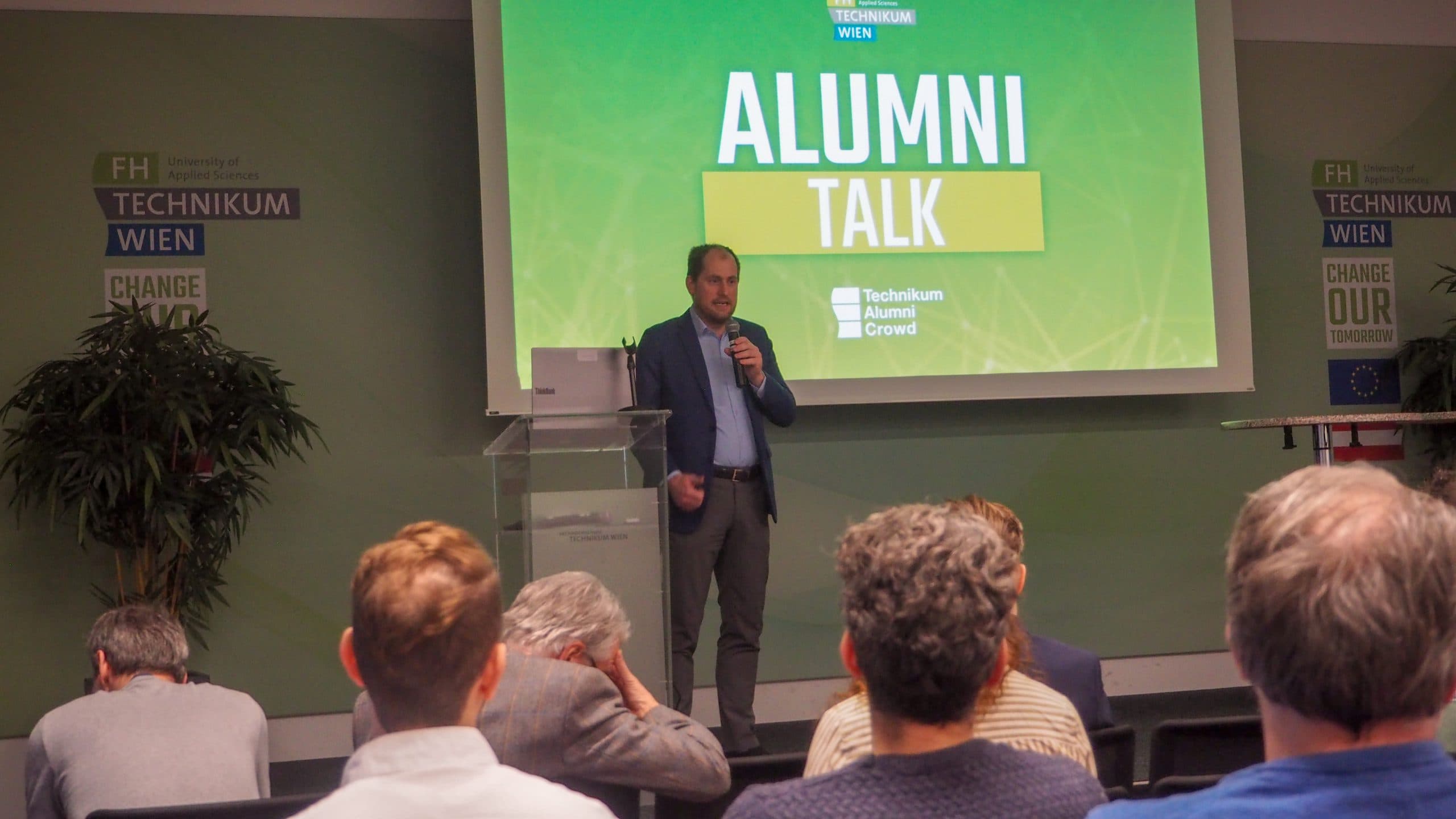 Ein Mann steht auf der Bühne und spricht bei einer ALUMNI TALK-Veranstaltung in ein Mikrofon, während mehrere Personen sitzen und zusehen. Ein großer grüner Bildschirm hinter ihm zeigt den Veranstaltungsnamen und das Logo der FH Technikum Wien.