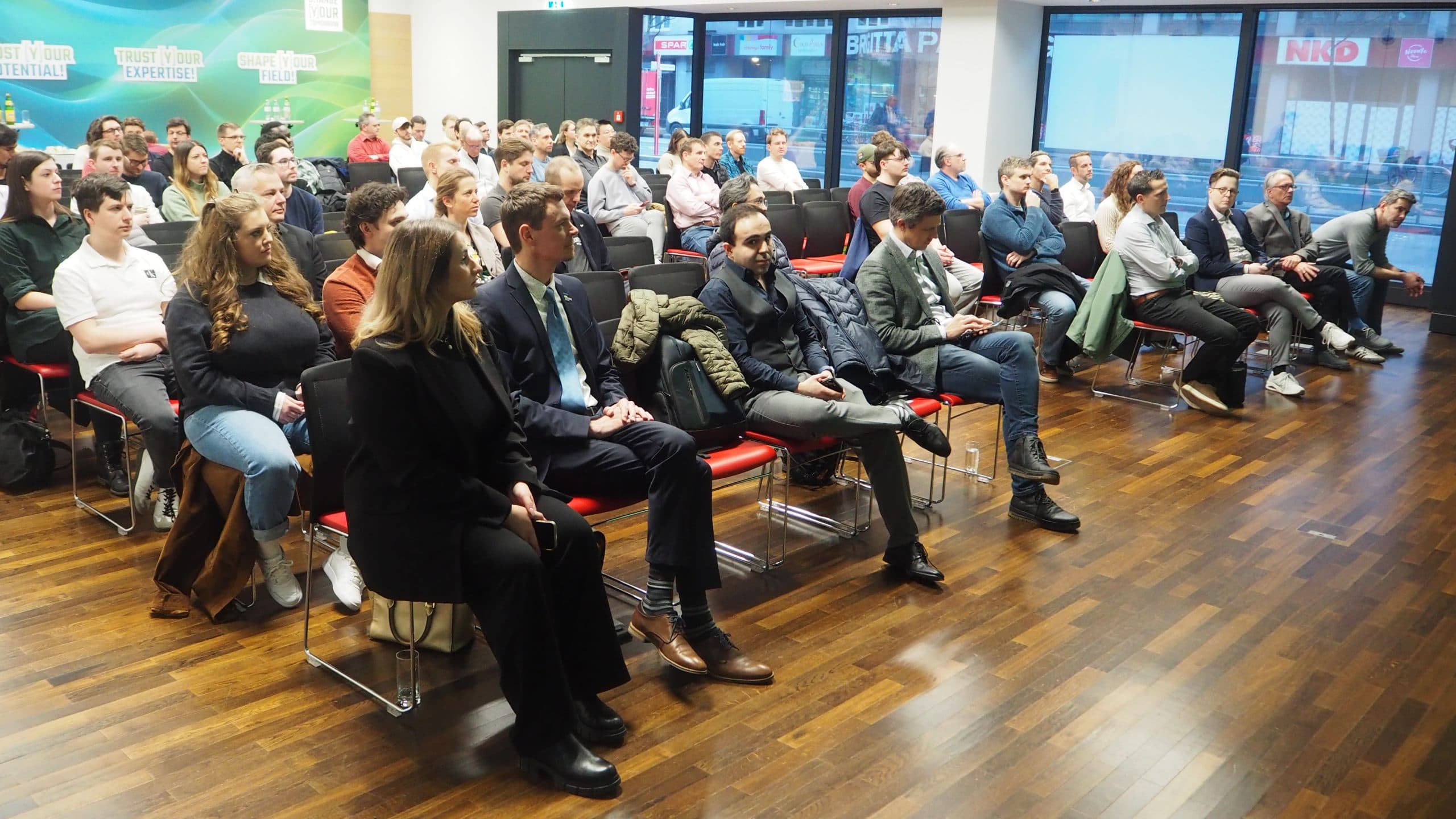 Eine Gruppe von Personen sitzt in Stuhlreihen mit Blickrichtung nach vorne in einem hellen Raum mit Holzfußboden und hört aufmerksam einer Präsentation oder einem Redner zu. Die meisten Teilnehmer scheinen engagiert zu sein, einige machen sich Notizen oder schauen auf Geräte.