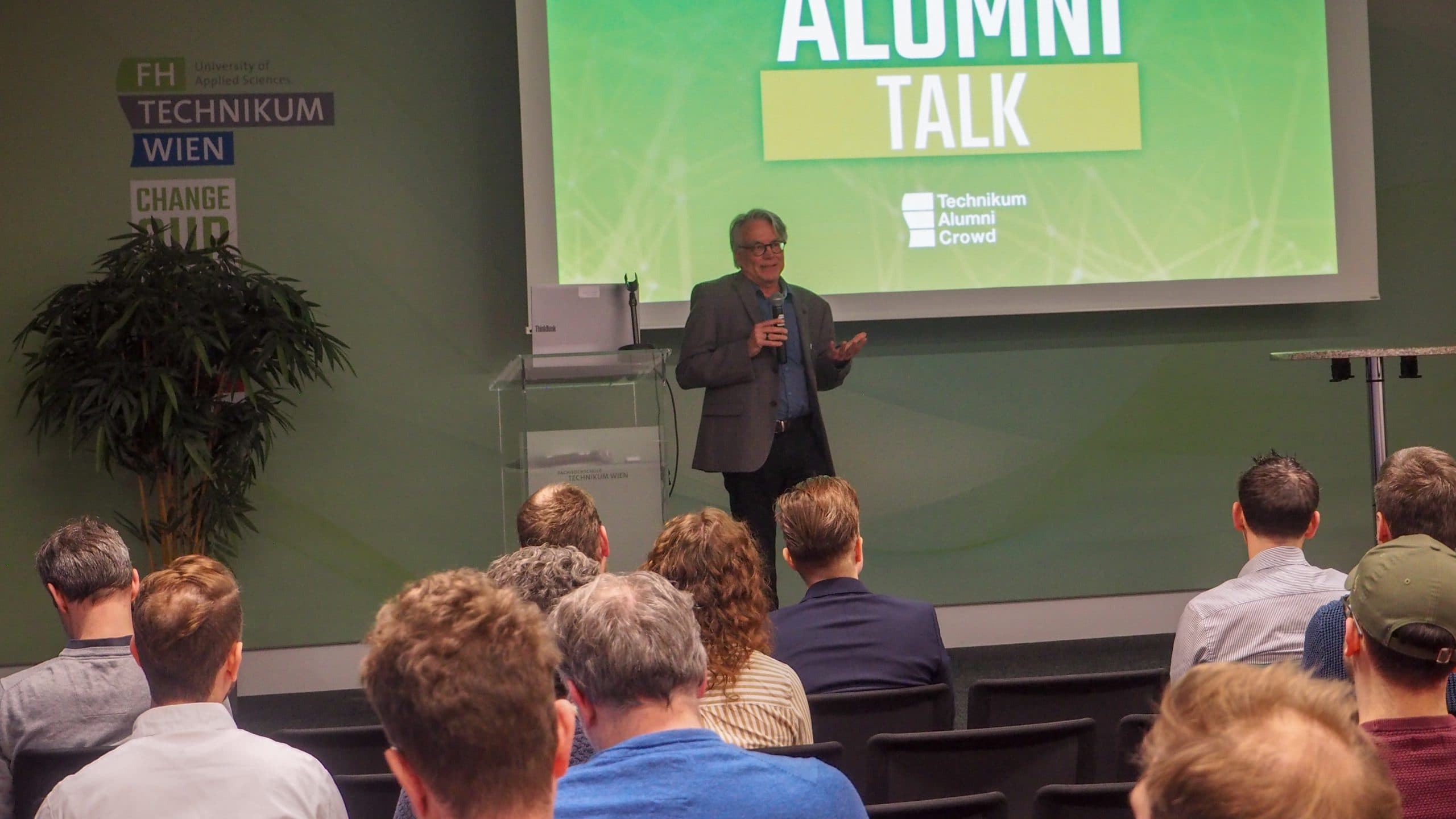 Ein Mann im Anzug spricht auf der Bühne zu den Zuhörern einer Veranstaltung namens 'Alumni Talk' an der FH Technikum Wien, hinter ihm eine grüne Präsentationsfolie und in Reihen sitzende Zuhörer.