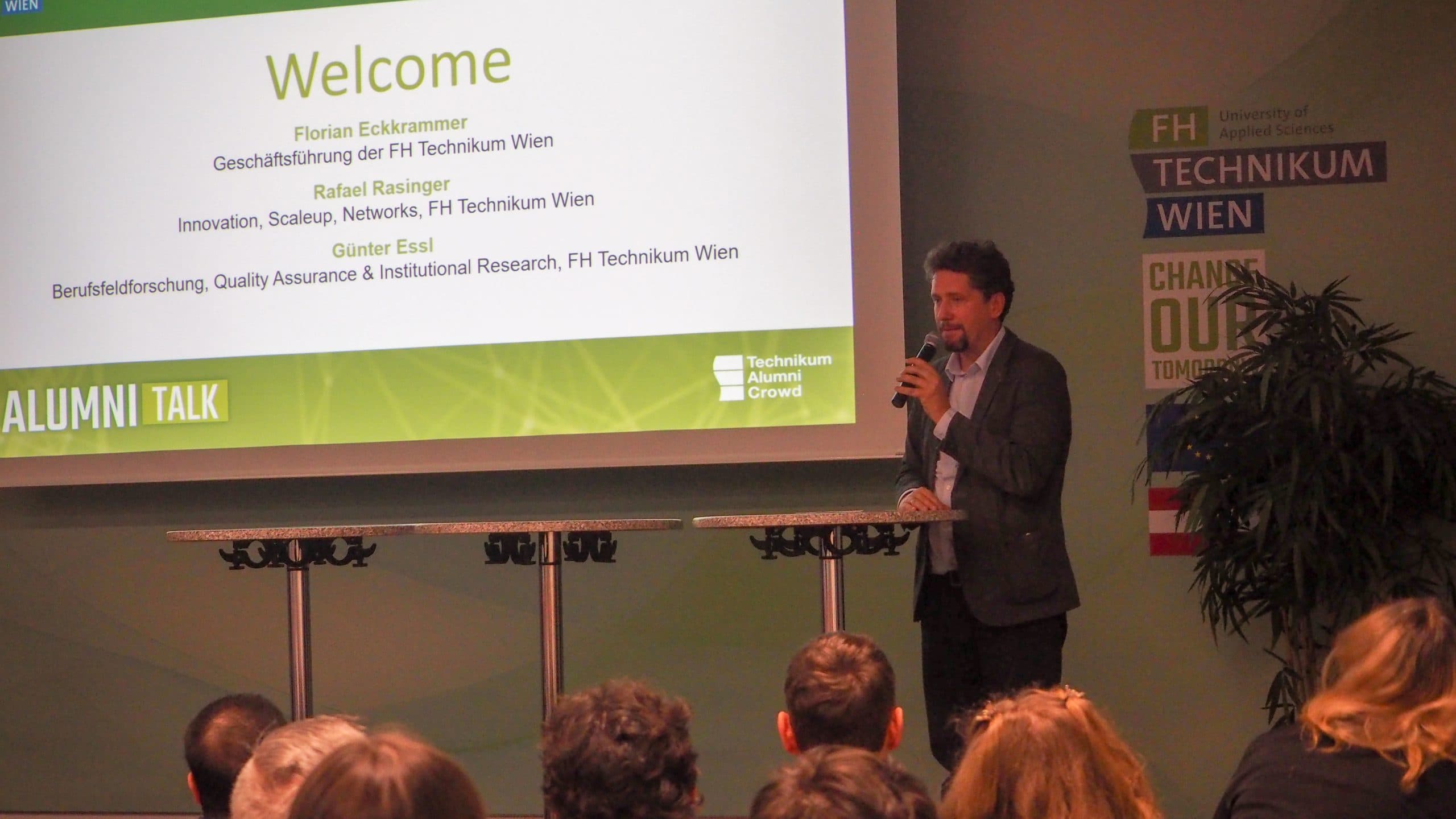 Ein Mann steht an einem Podium und spricht zu einem Publikum in einem Hörsaal. Eine projizierte Folie hinter ihm begrüßt die TeilnehmerInnen und listet die ReferentInnen für einen FH Technikum Wien Alumni Talk auf.