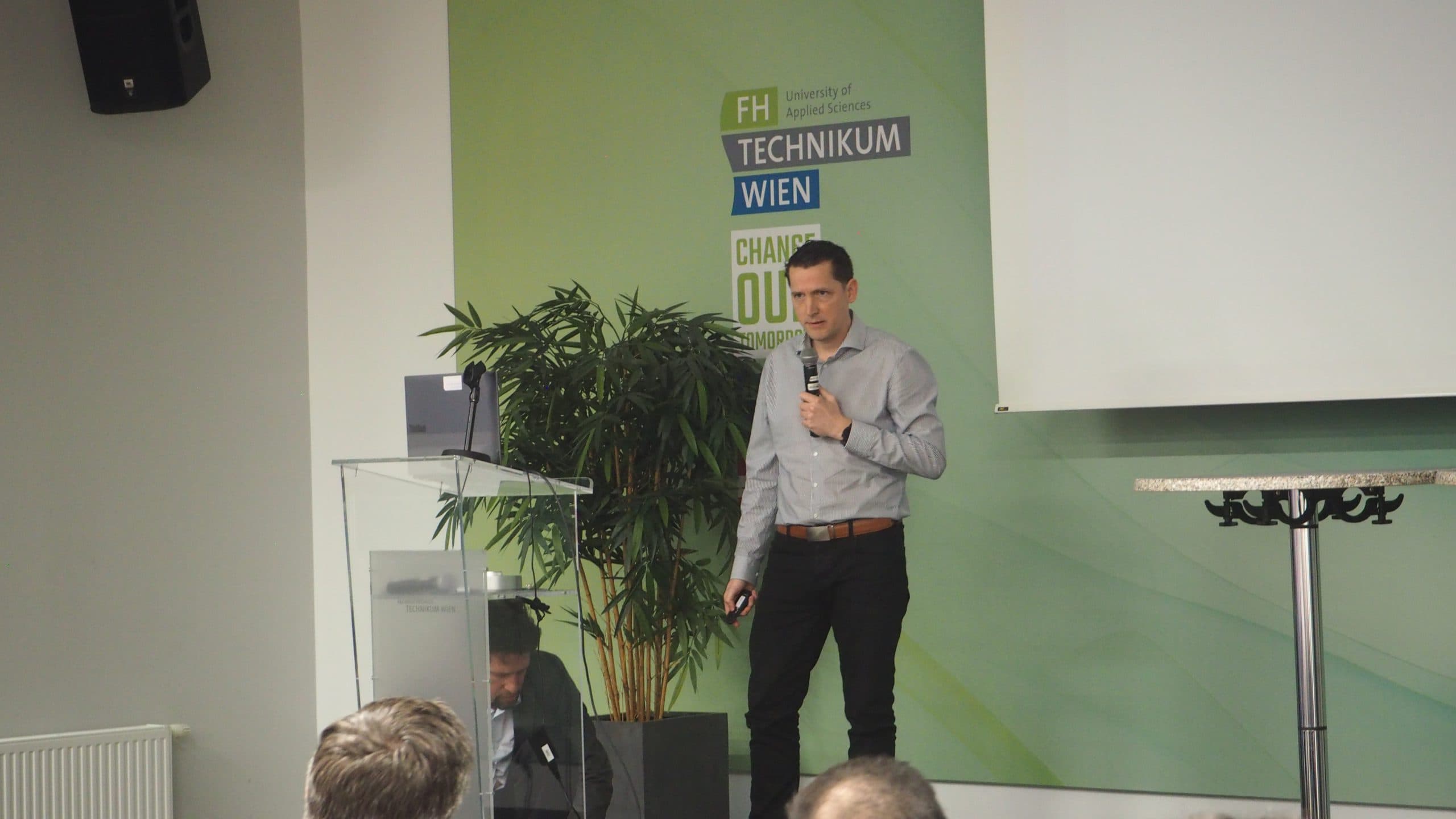 Ein Mann in einem hellgrauen Hemd steht auf der Bühne, hält ein Mikrofon und präsentiert vor einem Podium und einer grünen Wand mit der Aufschrift Technikum Wien. Im Vordergrund sind Zuhörer zu sehen.