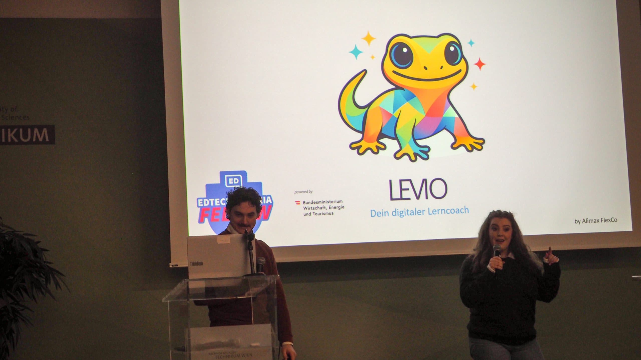 Zwei Personen präsentieren vor einer Projektionsfläche, auf der ein bunter Cartoon-Gecko und der Text 'LEMO Dein digitaler Lerncoach' zu sehen ist. Eine Person spricht an einem Podium, die andere gestikuliert neben ihr.