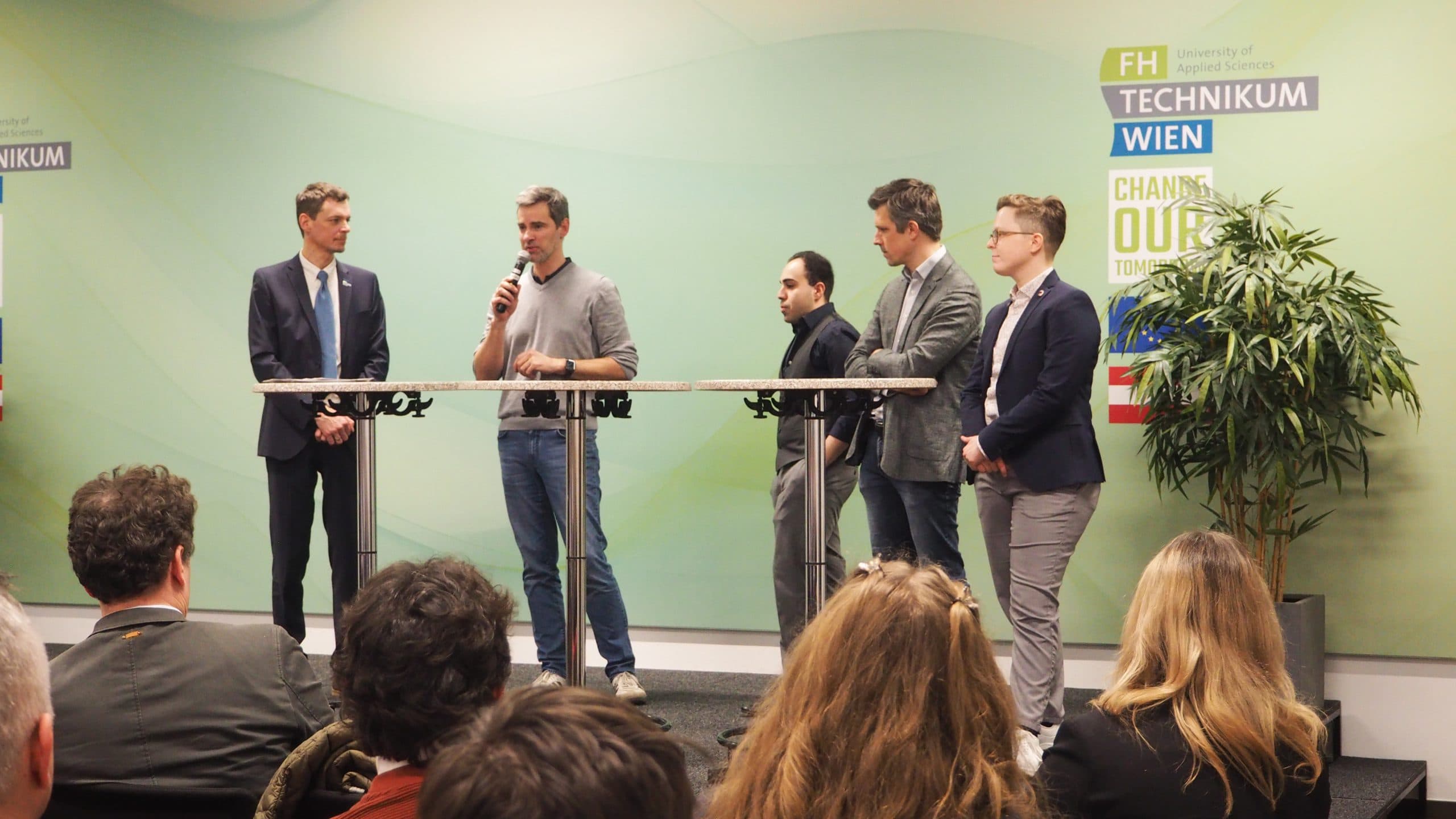 Fünf Männer stehen auf einer Bühne vor einem Publikum an der FH Technikum Wien. Ein Mann spricht in ein Mikrofon, während die anderen zuhören. Im Hintergrund sind Transparente und eine Pflanze zu sehen.