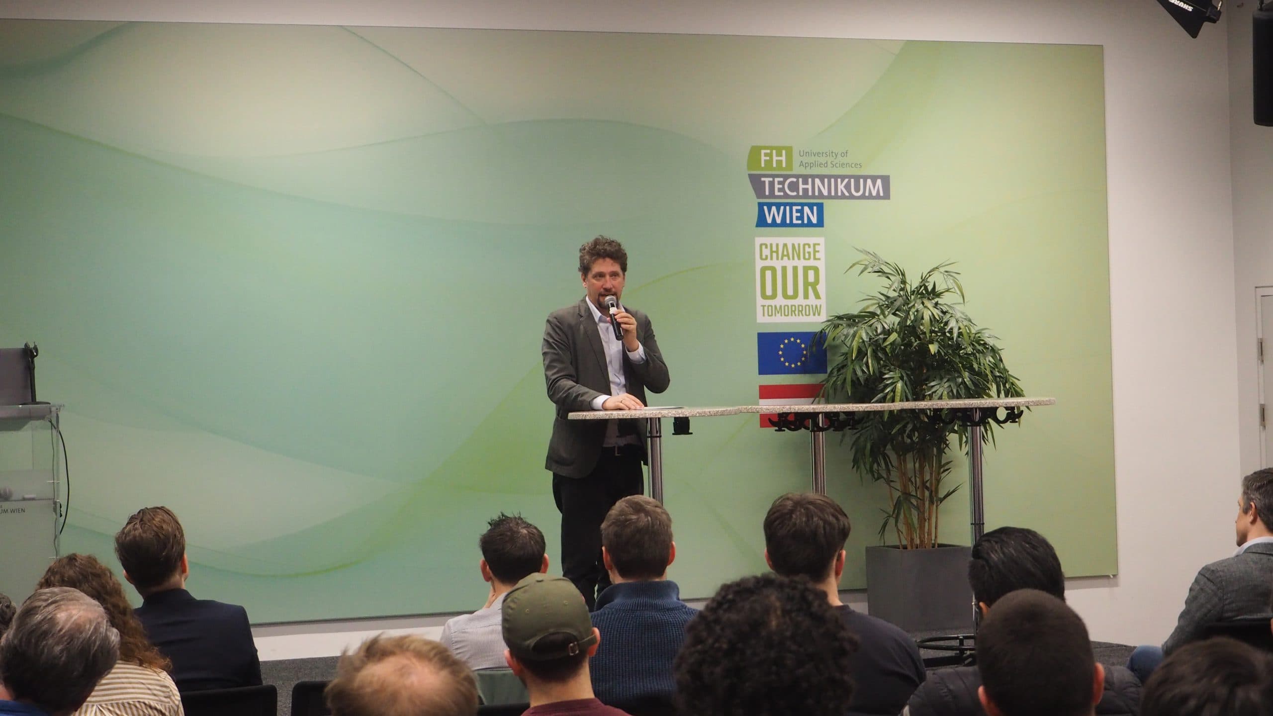 Ein Mann im Anzug spricht auf einem Podium vor einem Publikum in ein Mikrofon. Hinter ihm sind Schilder für die FH Technikum Wien und Change Our Tomorrow sowie EU- und Österreich-Flaggen zu sehen. Eine Topfpflanze steht neben dem Podium.