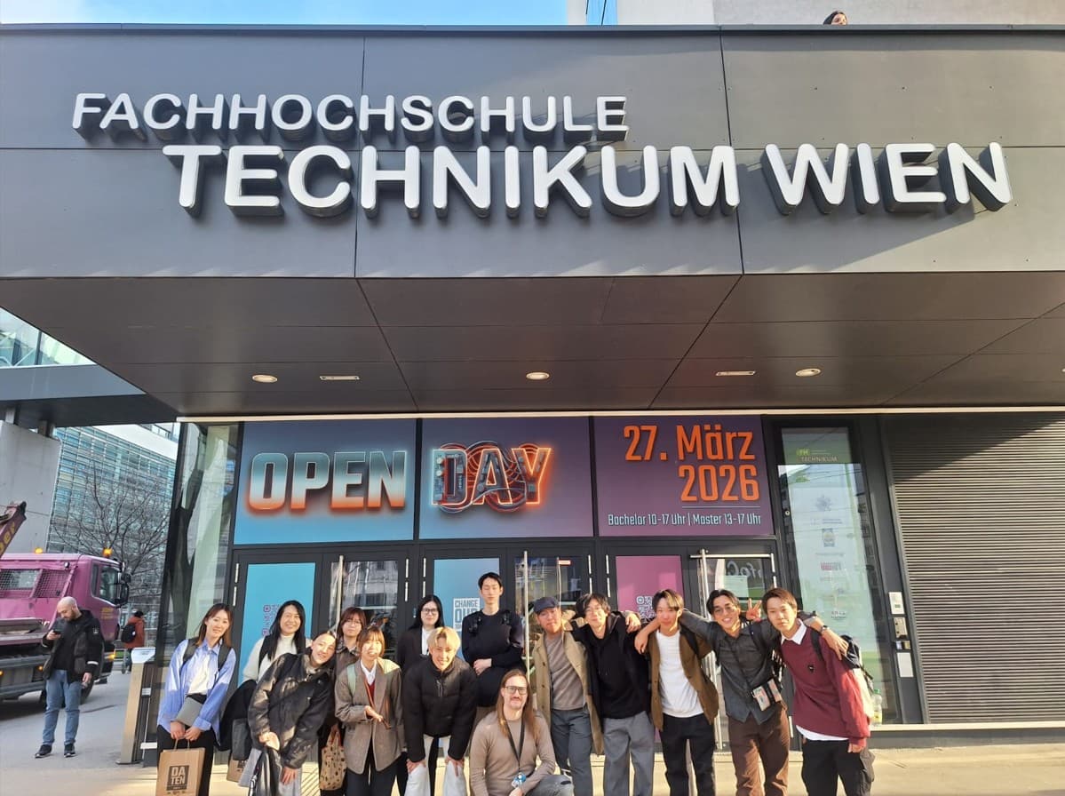 Eine Gruppe von dreizehn lächelnden Menschen posiert vor dem Gebäude der Fachhochschule Technikum Wien und steht unter einem großen Schild. Plakate an den Fenstern werben für einen Tag der offenen Tür am 27. März 2026.