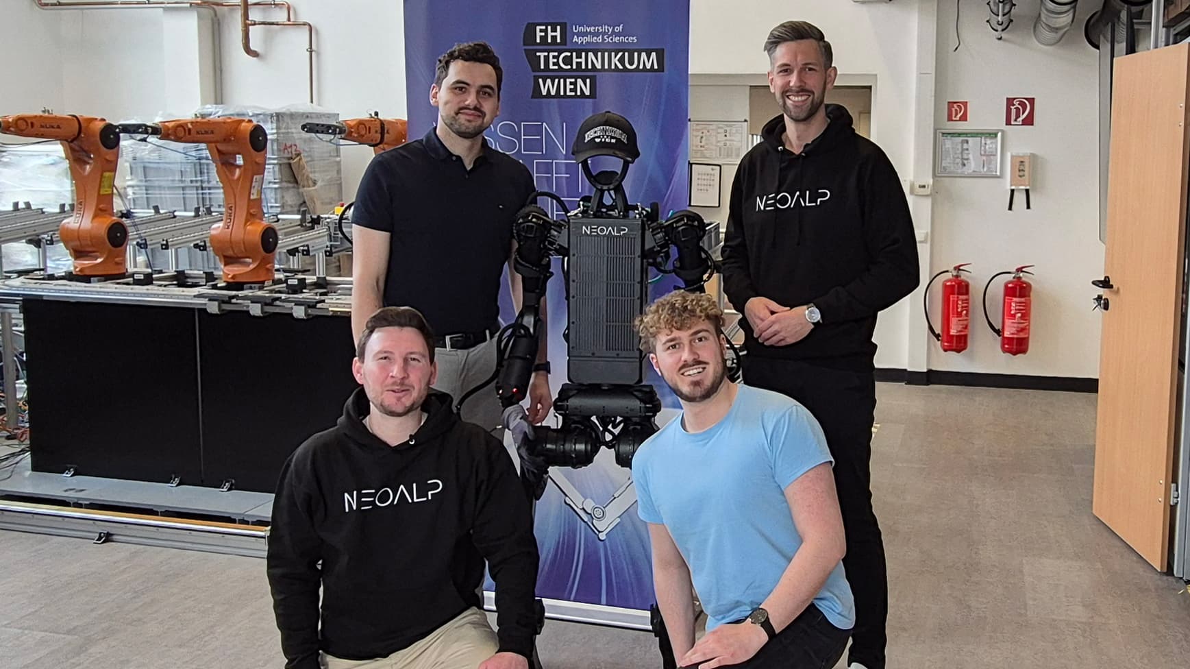 Vier Männer posieren mit einem humanoiden Roboter vor einem Banner der FH Technikum Wien in einem modernen Labor. Zwei Männer tragen NEONALP Kapuzenpullis. Im Hintergrund sind Industrieroboterarme und Feuerlöscher zu sehen.