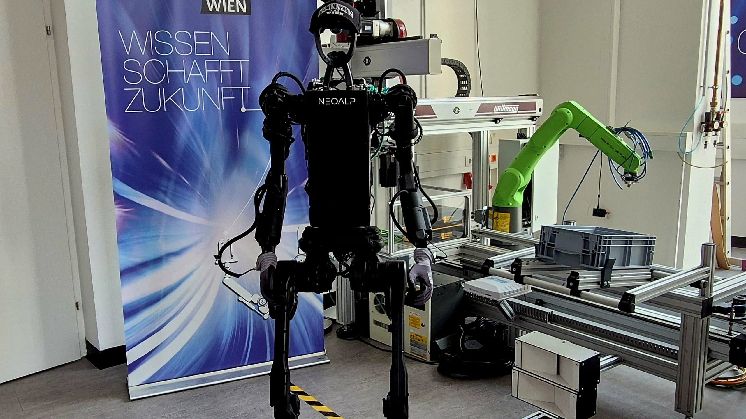 Ein humanoider Roboter steht in einem technischen Labor neben Maschinen und Geräten. Im Hintergrund ist ein blaues Banner mit der Aufschrift Wissen Schafft Zukunft zu sehen.