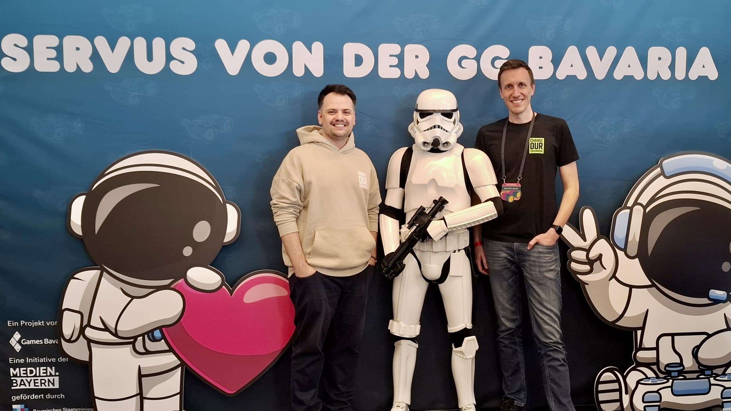 Zwei Männer stehen zu beiden Seiten einer Person im Stormtrooper-Kostüm, die vor einem blauen Hintergrund mit "Servus von der GG Bavaria" und Cartoon-Astronautenfiguren posiert und lächelt.