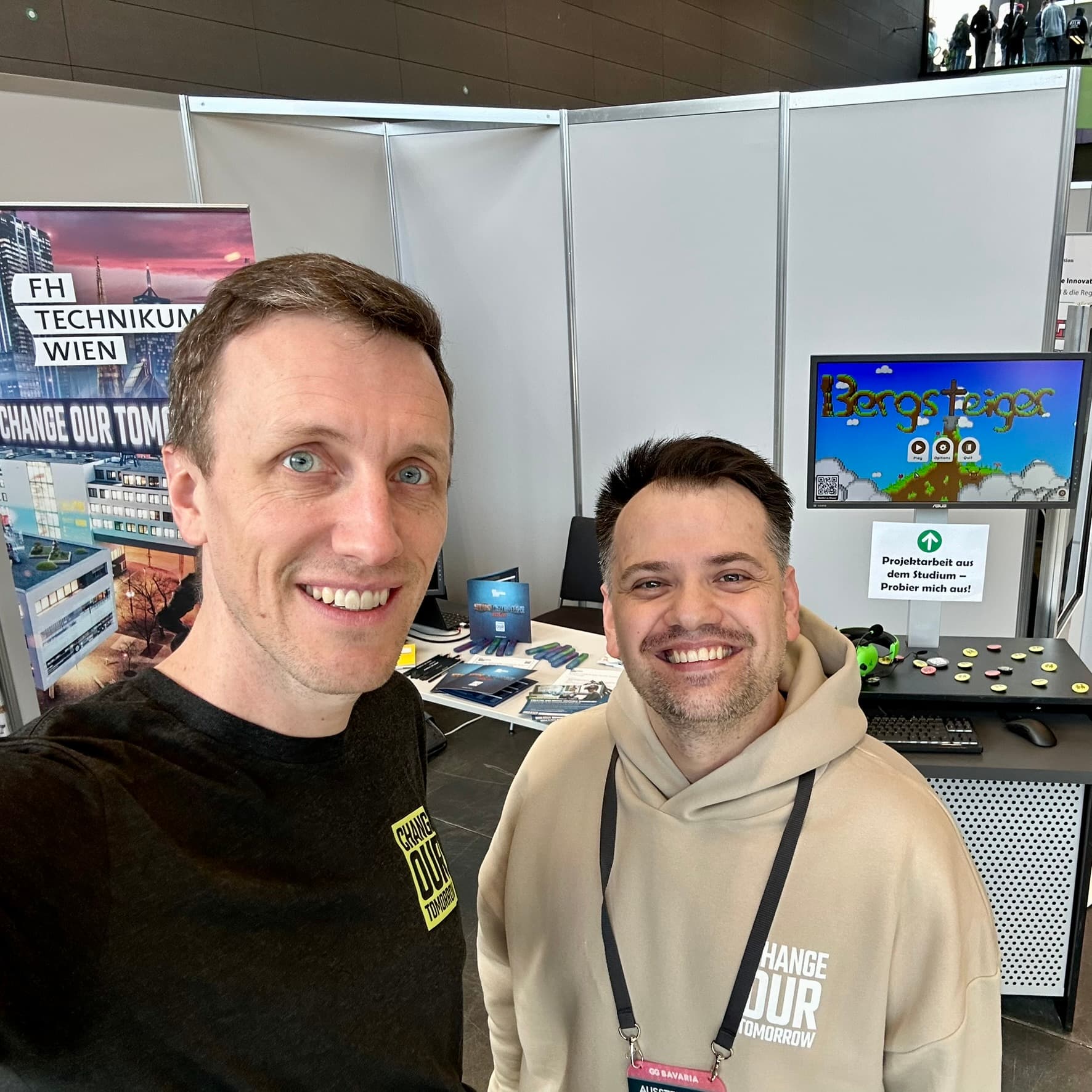 Zwei lächelnde Männer stehen bei einer Veranstaltung vor einem Stand. Hinter ihnen sind Banner, ein Monitor mit dem Spiel 'Bergsteiger' und Informationsmaterial auf Tischen zu sehen. Der Stand wirbt für die FH Technikum Wien.