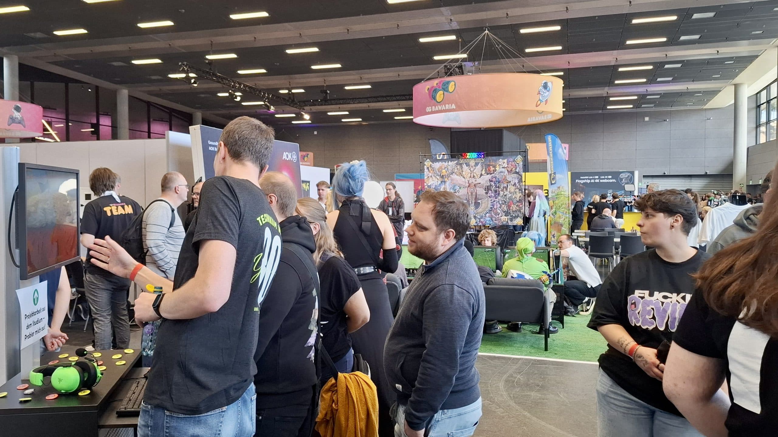 Eine belebte Kongresshalle mit Menschen, die an verschiedenen Ständen interagieren, Spiele spielen und Kontakte knüpfen. Bunte Banner und Displays sind zu sehen, und die Teilnehmer tragen legere Kleidung, einige mit Marken-T-Shirts.