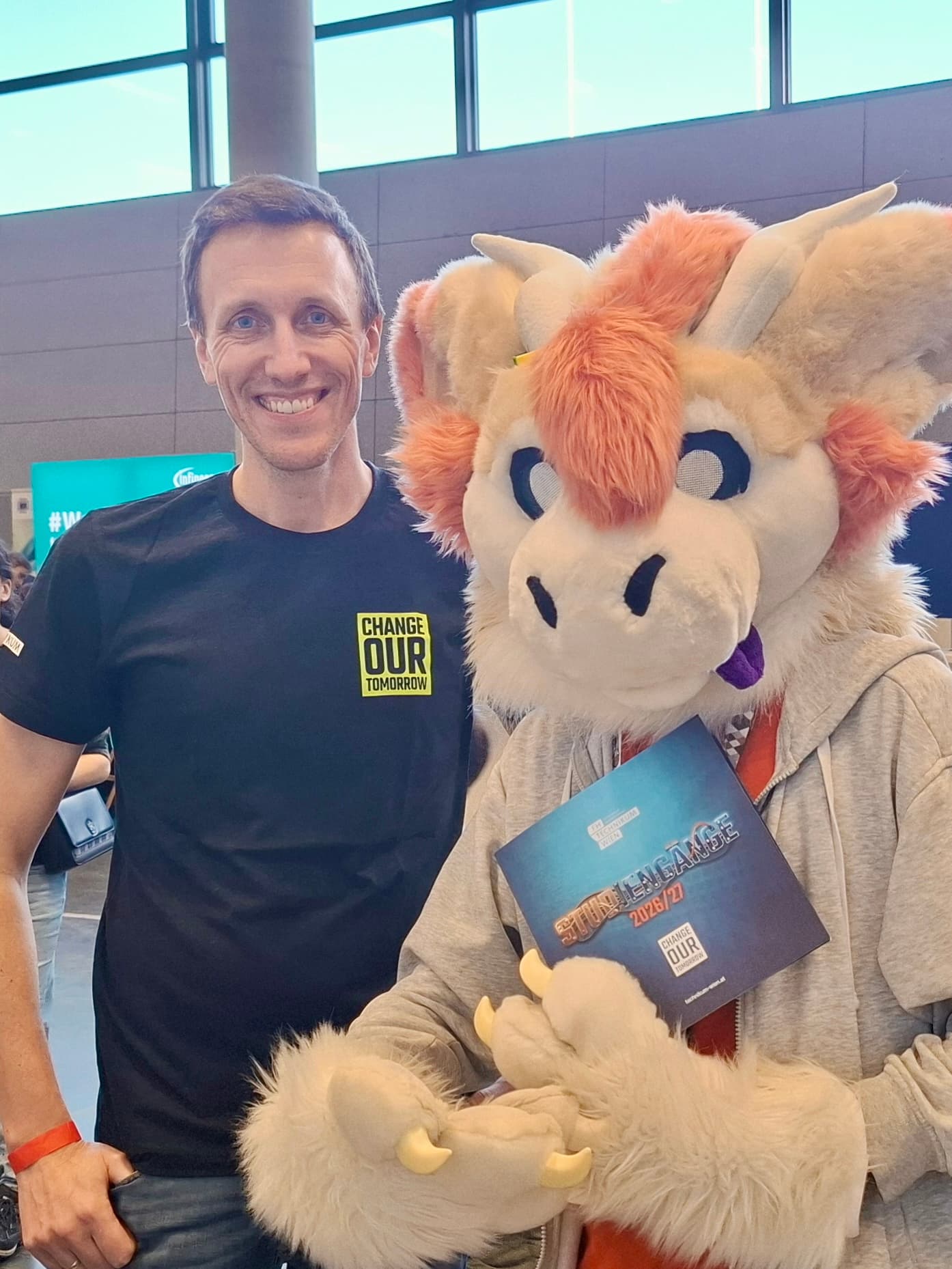 Eine Person in einem schwarzen Change Our Tomorrow-T-Shirt steht neben einer Person in einem pelzigen Drachenkostüm, die bei einer Indoor-Veranstaltung ein Programm für die Sommerspiele 2023 hält. Beide lächeln in die Kamera.