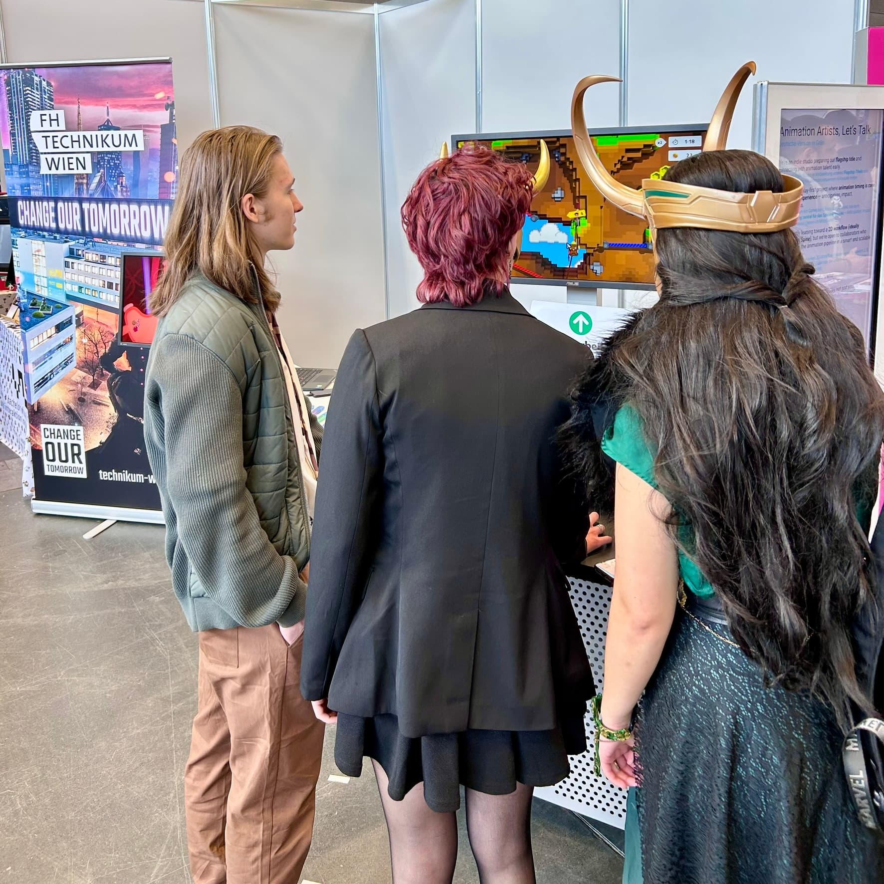Drei Personen stehen an einem Messestand vor einem Bildschirm, auf dem ein buntes Spiel gezeigt wird. Eine Person trägt einen gehörnten Helm und ein grünes Outfit. Im Hintergrund sind Plakate und Flugblätter zu sehen, die für die FH Technikum Wien werben.