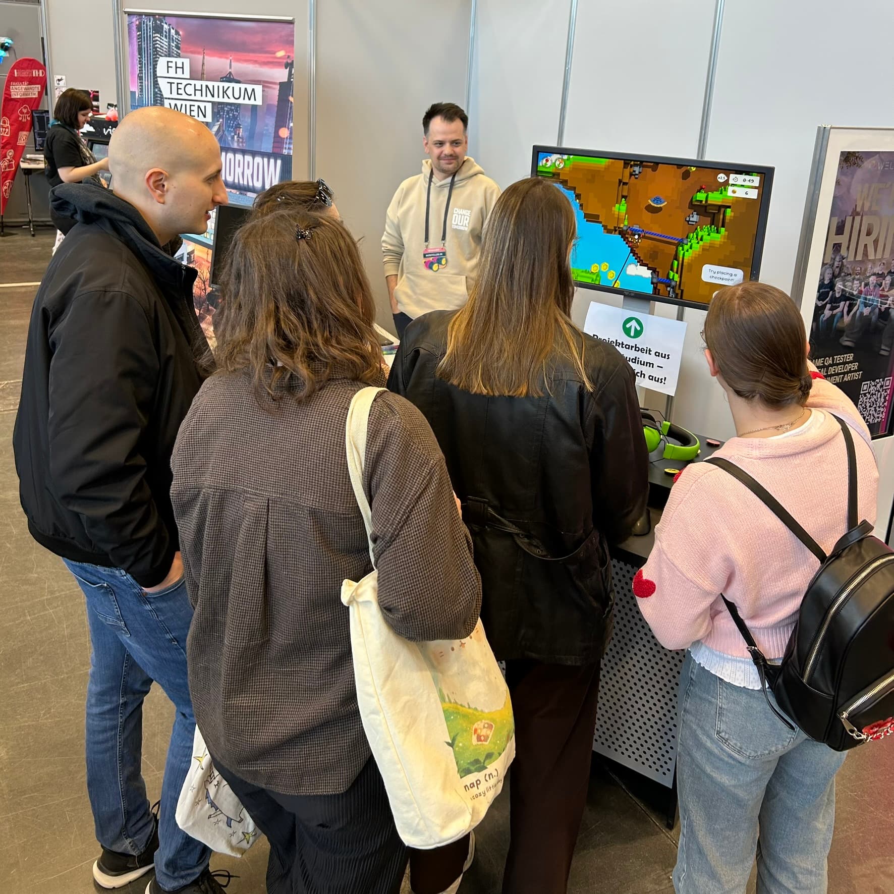 Eine Gruppe von Menschen steht vor einem Ausstellungsstand und beobachtet, wie ein Mann ein Spiel oder einen interaktiven Bildschirm vorführt. Im Hintergrund sind Plakate und Banner der Veranstaltung zu sehen.