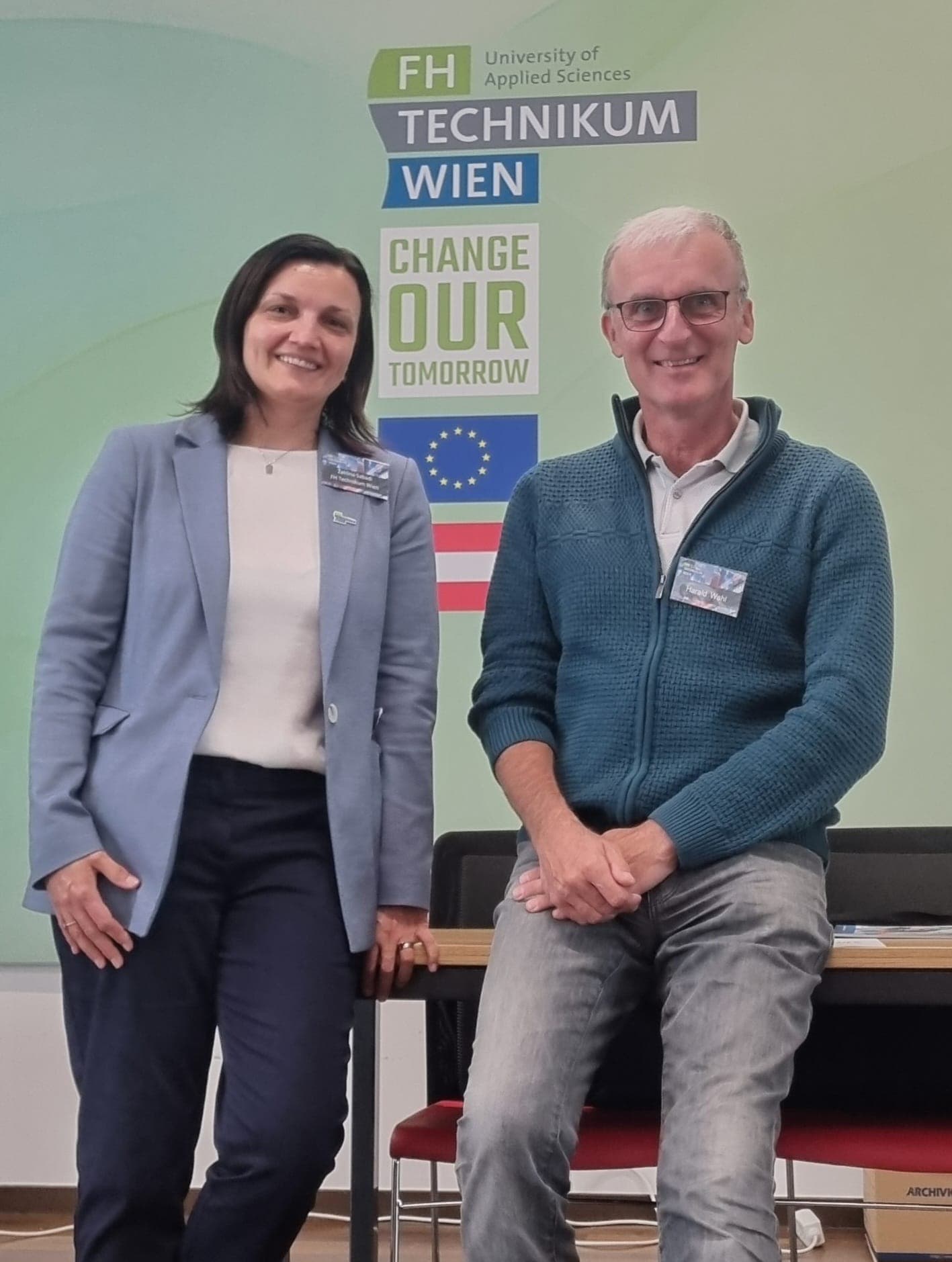 Zwei Personen lächeln und posieren vor einer Wand mit Logos der FH Technikum Wien und dem Slogan Change Our Tomorrow. Die eine sitzt auf einem Tisch, die andere steht daneben.