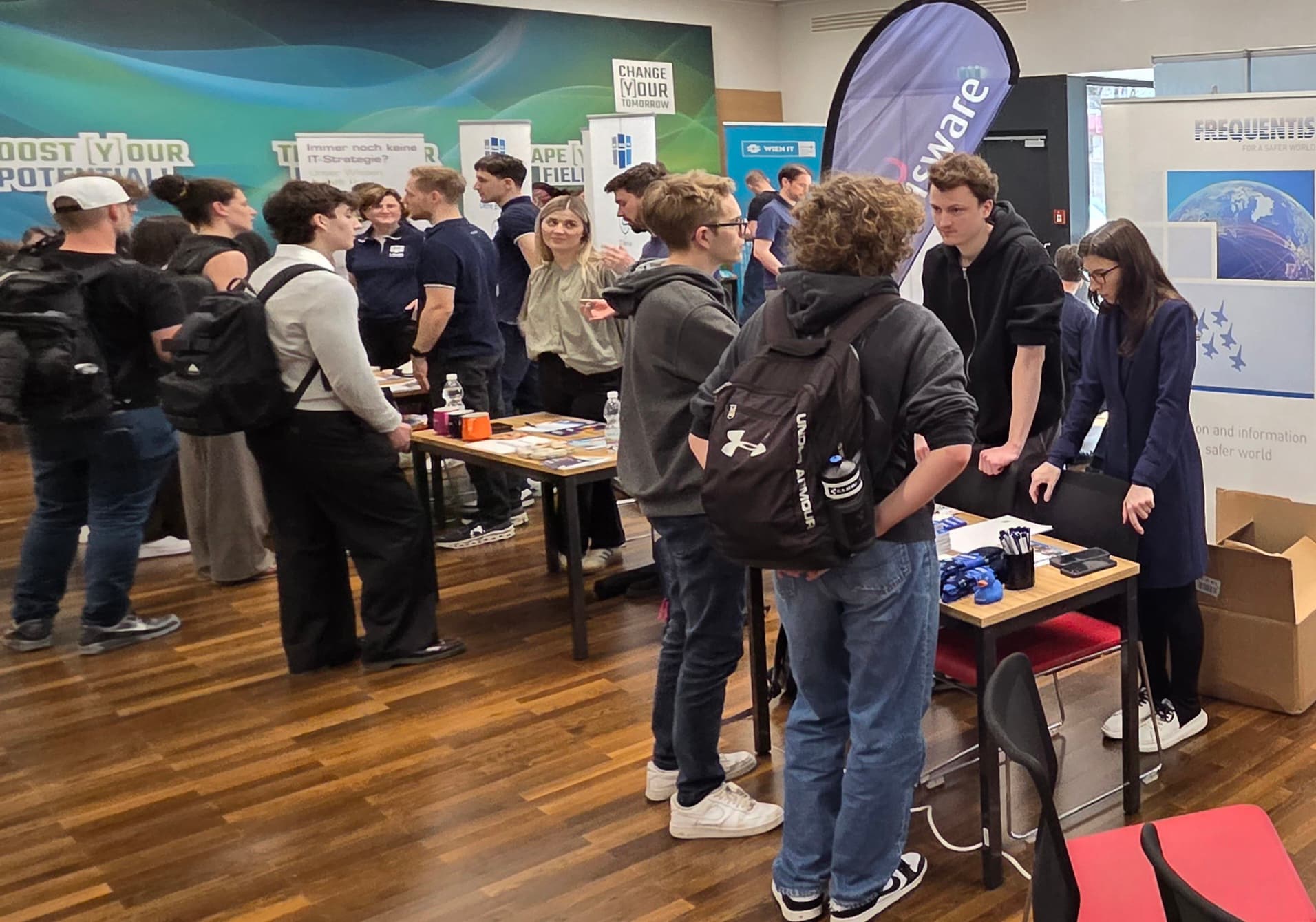 Eine Gruppe von Studenten unterhält sich mit Unternehmensvertretern auf einer belebten Karrieremesse in einer Halle, umgeben von Werbeständen, Bannern und Informationsmaterial.
