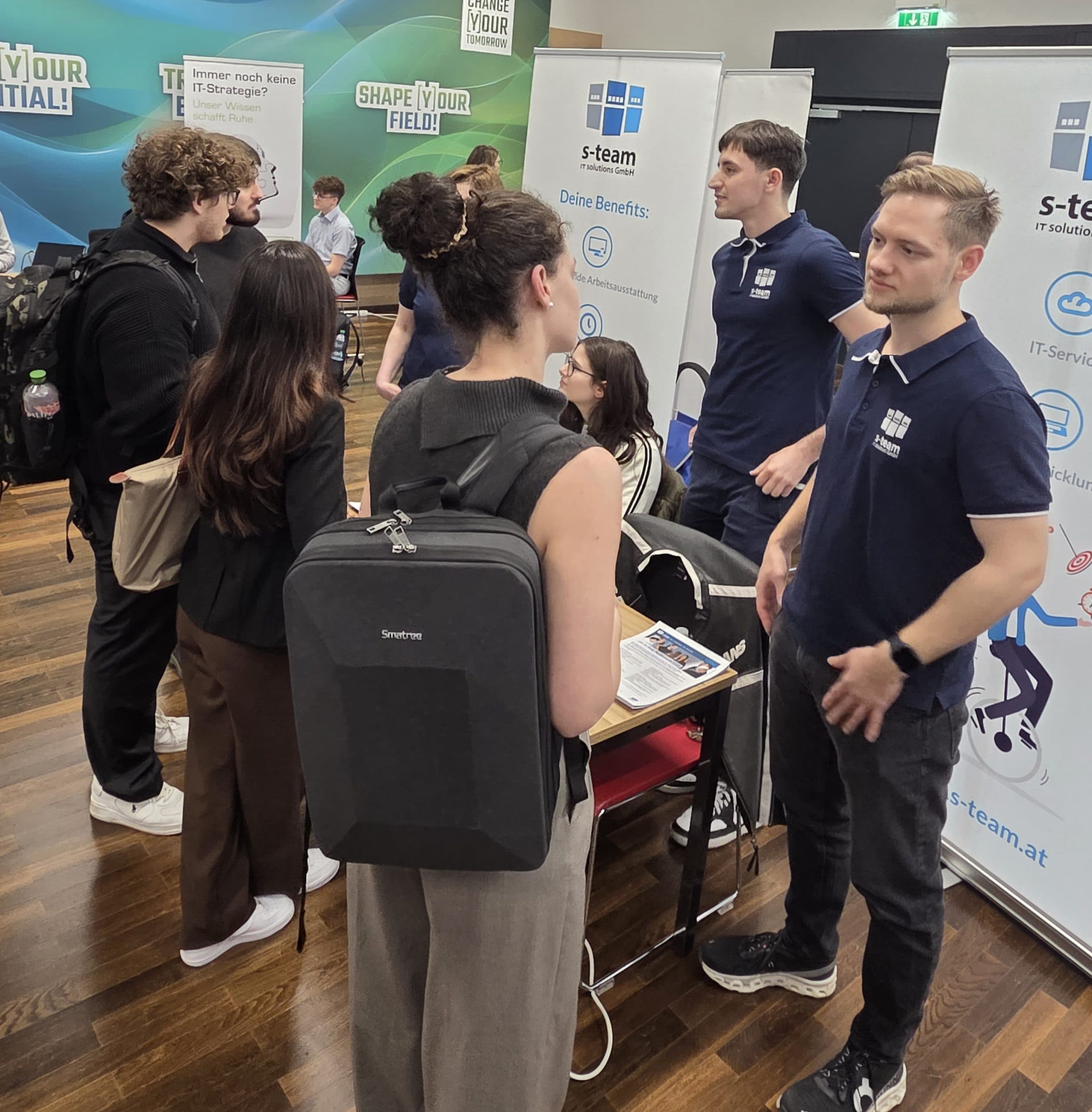 Menschen unterhalten sich an einem Stand auf einer Karrieremesse mit 's-team'-Bannern. Zwei Männer in marineblauen Polos unterhalten sich mit einer Frau mit einem schwarzen Rucksack. Andere treffen sich zum Networking in einem modernen Raum mit Holzböden und Informationsdisplays.