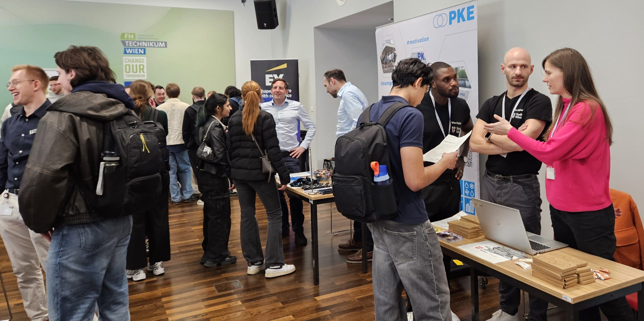 Eine Gruppe von Menschen tauscht sich auf einer überdachten Karrieremesse aus. Einige stehen an Tischen mit Laptops, Displays und Broschüren, während andere sich unterhalten, Kontakte knüpfen oder Materialien prüfen. Im Hintergrund sind Banner und Informationsschilder zu sehen.