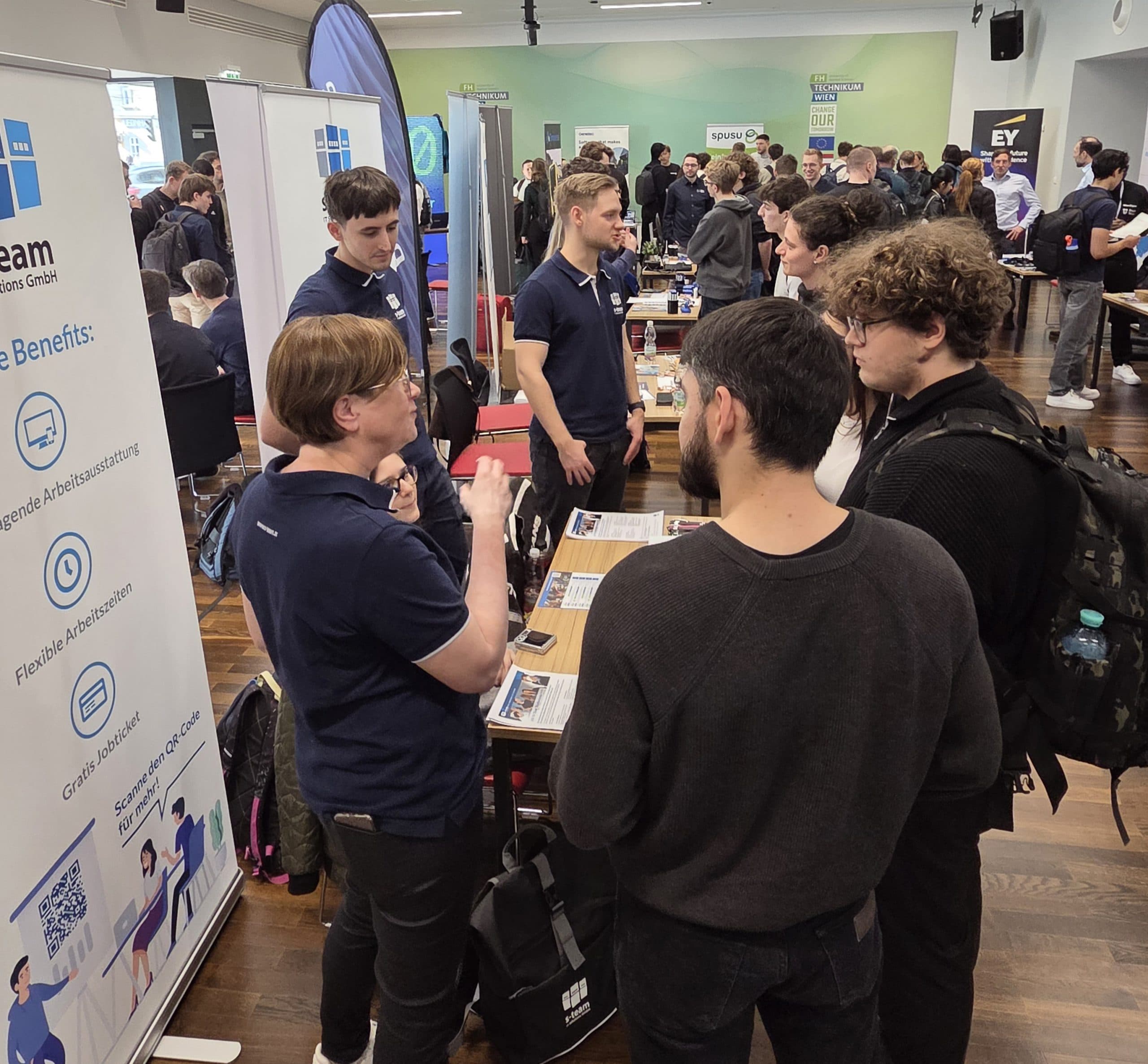 Menschen besuchen eine Jobmesse in einem Gebäude und unterhalten sich in Gruppen. Ein Team an einem Stand in marineblauen Hemden spricht mit den Besuchern. Banner und Werbematerialien sind zu sehen, und der Raum ist voll mit Besuchern und Ausstellern.