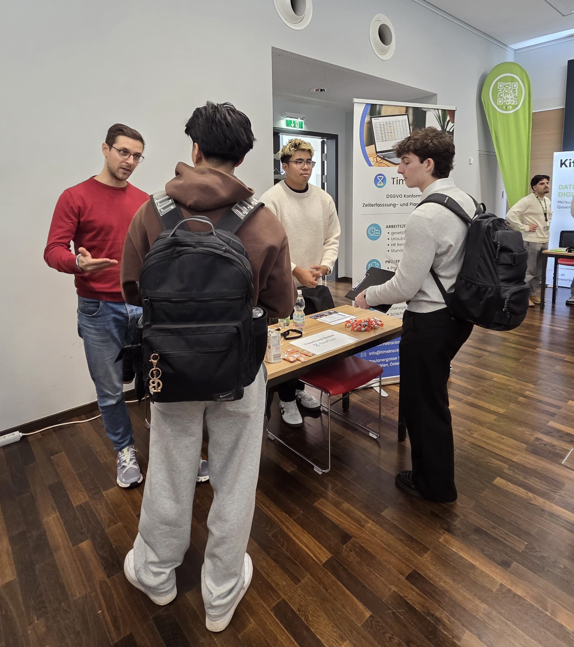 Drei junge Männer mit Rucksäcken unterhalten sich mit zwei Vertretern an einem Stand mit Informationsmaterial bei einer Indoor-Veranstaltung. Der Stand hat Banner, einen Laptop und Werbeartikel auf dem Tisch.