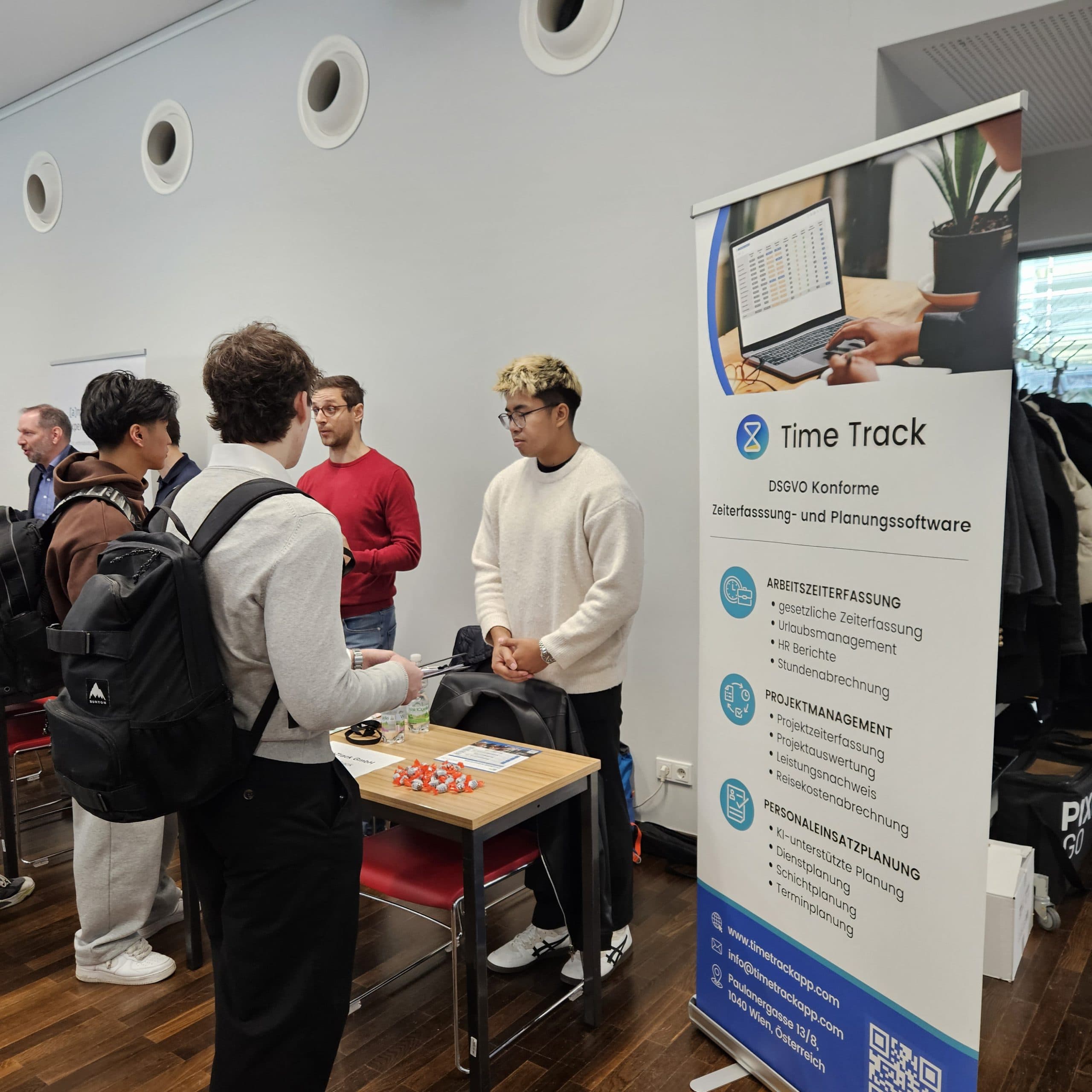 Eine Gruppe junger Männer unterhält sich bei einer Indoor-Veranstaltung neben einem Banner für die Software Time Track, auf der Funktionen wie Zeiterfassung und Projektmanagement aufgeführt sind. Ein Mann steht hinter einem Tisch mit Flyern und Werbeartikeln.