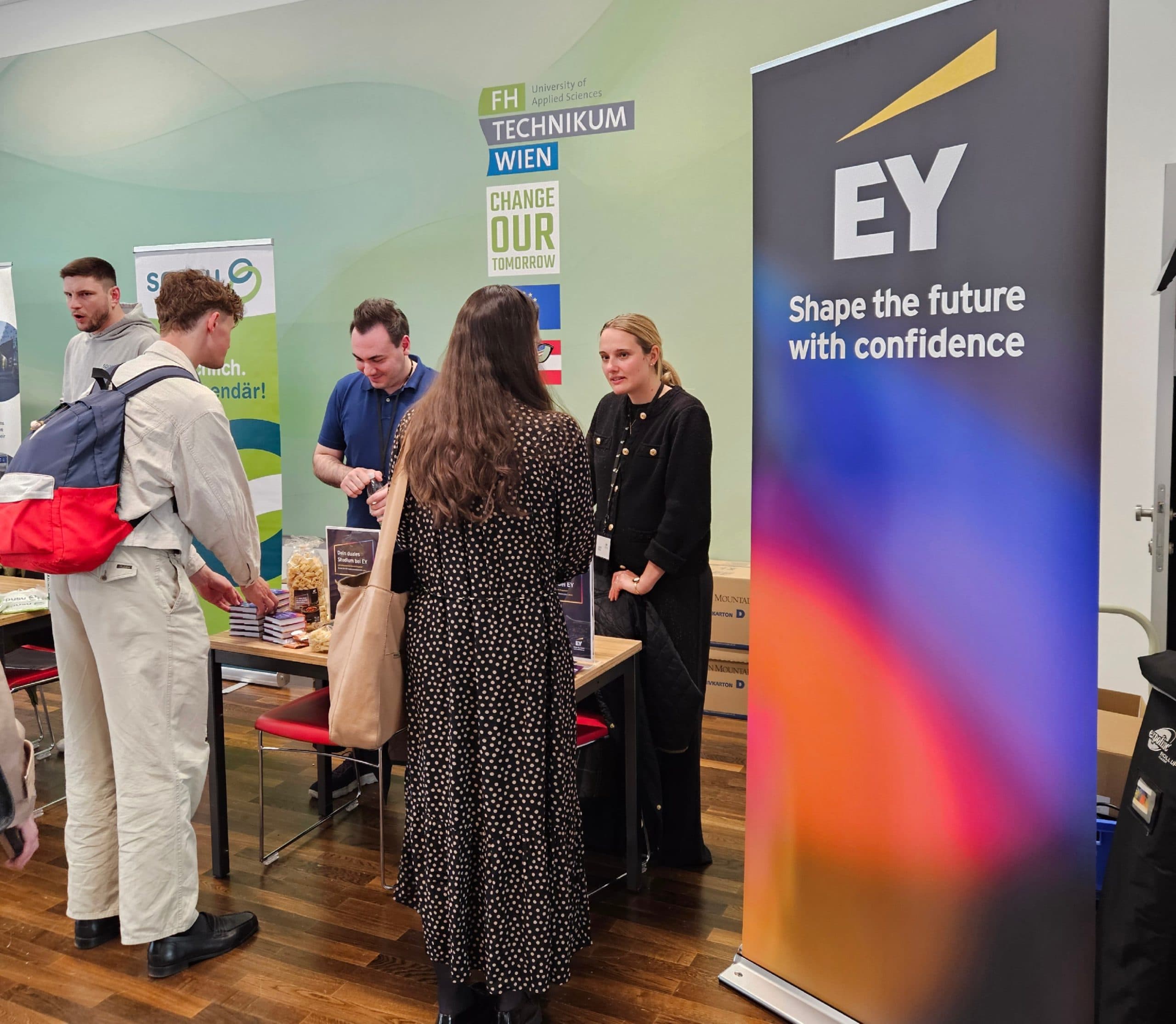 Eine Gruppe von Menschen unterhält sich auf einer Karrieremesse an einem Stand mit einem EY-Banner, auf dem 'Shape the future with confidence' steht. Im Hintergrund sind weitere Stände und Banner zu sehen.