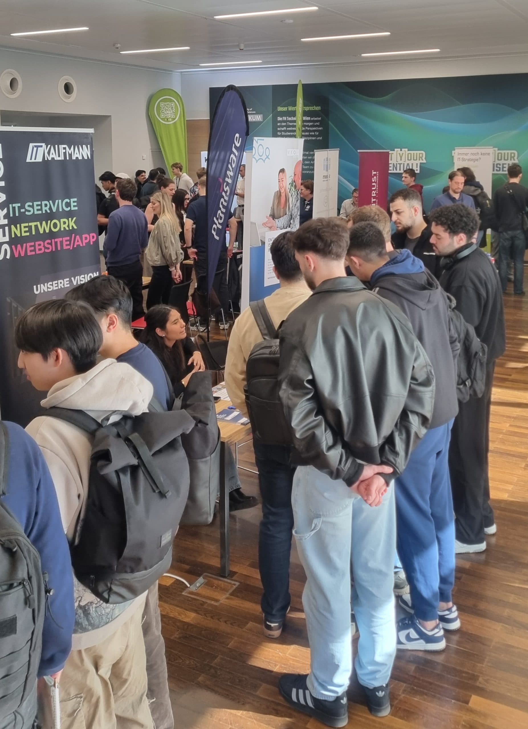 Eine Menschenmenge, viele mit Rucksäcken, steht Schlange und besucht Stände auf einer überdachten Berufsmesse oder Expo mit Bannern und Informationsdisplays.