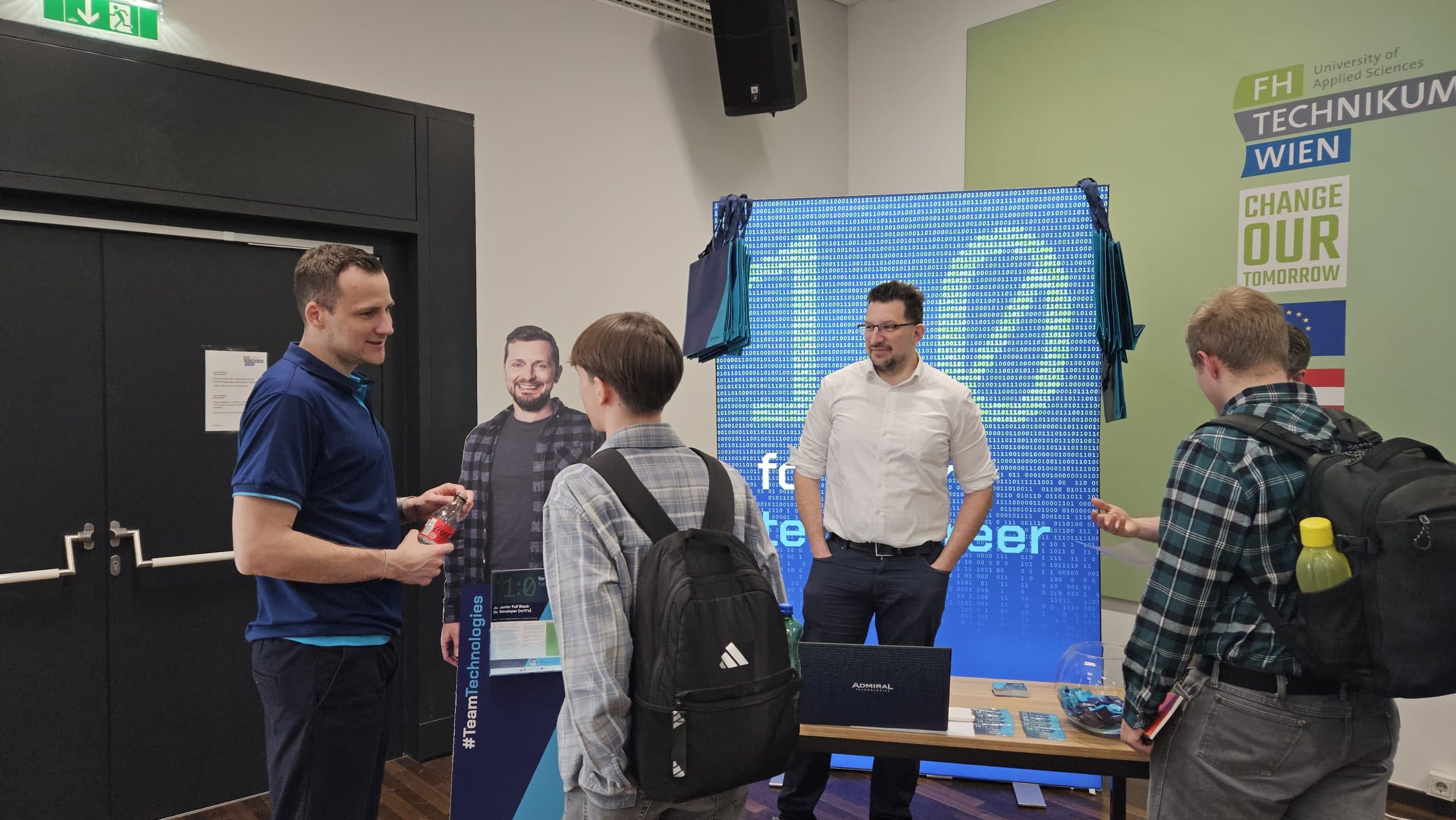Zwei Männer unterhalten sich mit zwei Studenten an einem Informationsstand der FH Technikum Wien. Der Stand zeigt Werbematerialien, einen Laptop und einen Pappausschnitt eines Mannes. Auf einem großen Schild steht 'Change Our Tomorrow'.
