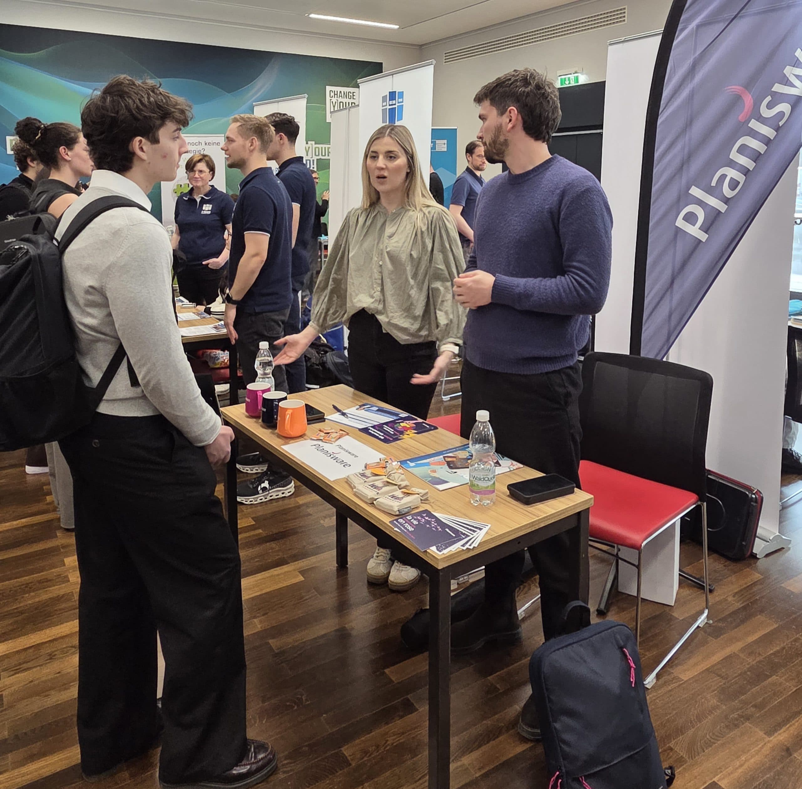 Drei Personen stehen während einer Karrieremesse an einem Stand von Planswell und unterhalten sich. Am Stand liegen Broschüren, Tassen und Wasserflaschen auf dem Tisch. Im Hintergrund sind andere Stände und Besucher zu sehen.