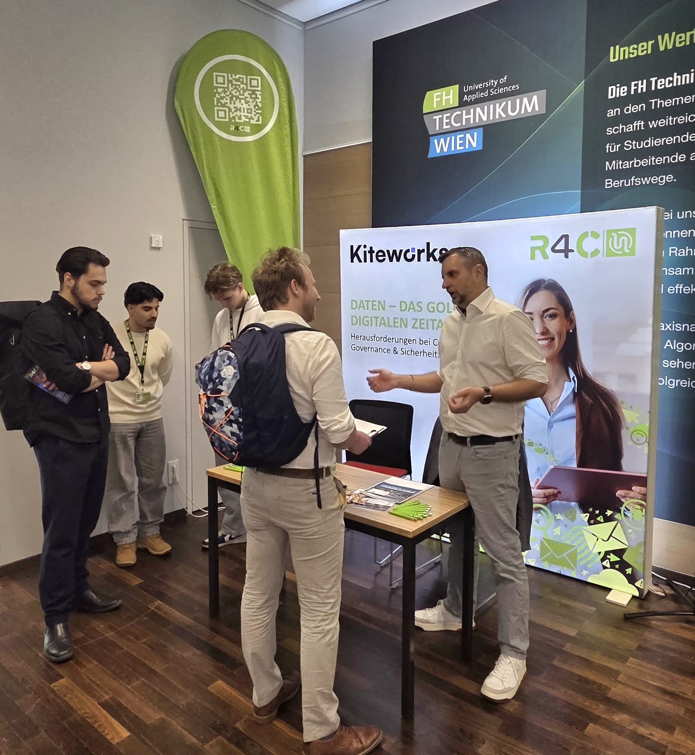 Vier Personen stehen an einem Informationsstand auf einer Berufsmesse. Eine Person am Stand überreicht einer Besucherin einen Flyer. Banner und ein Tisch mit Broschüren werben für die FH Technikum Wien und digitale Datenkarrieren.