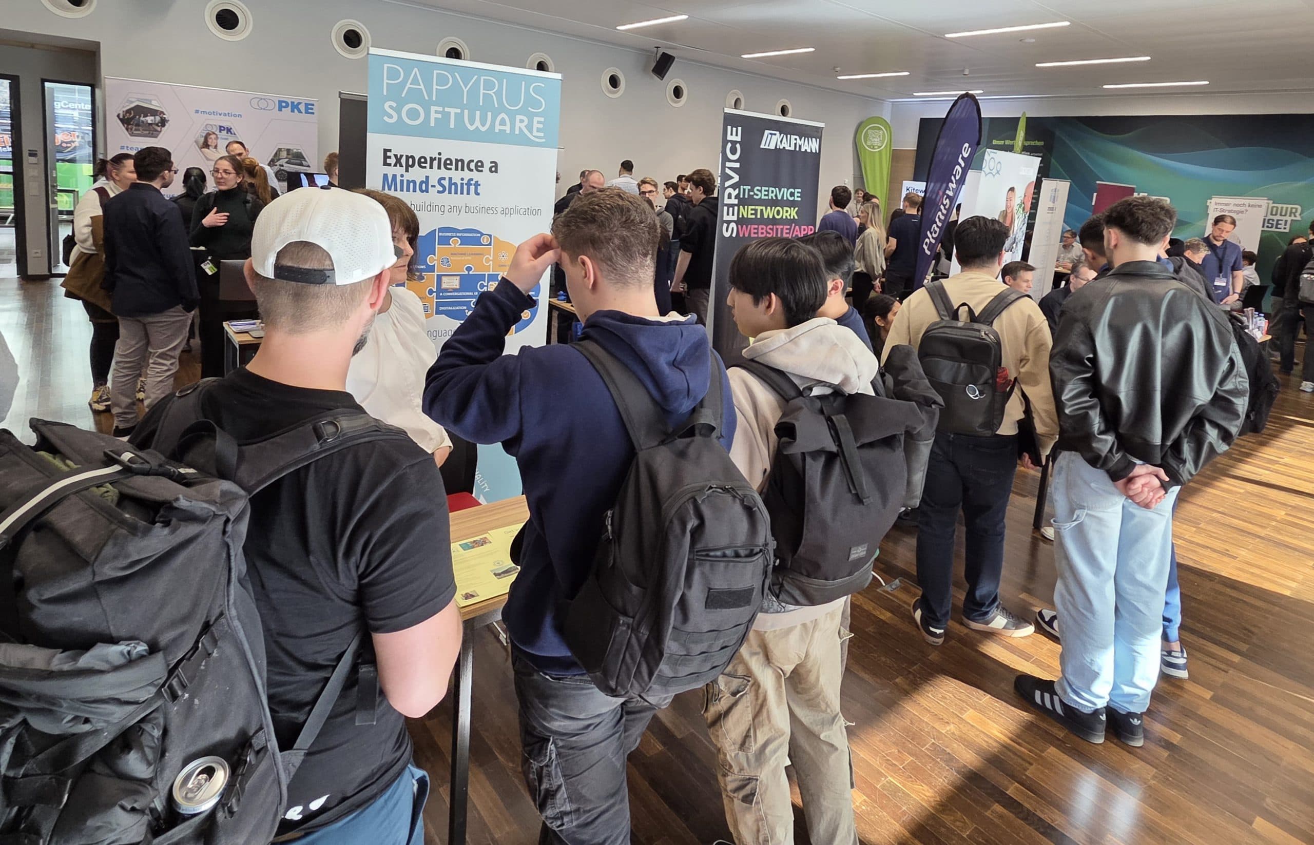 Eine Gruppe von Menschen, viele mit Rucksäcken, steht auf einer Jobmesse Schlange und besucht die Stände von Unternehmen mit Bannern und Displays in einem modernen, hell erleuchteten Raum mit Holzboden.