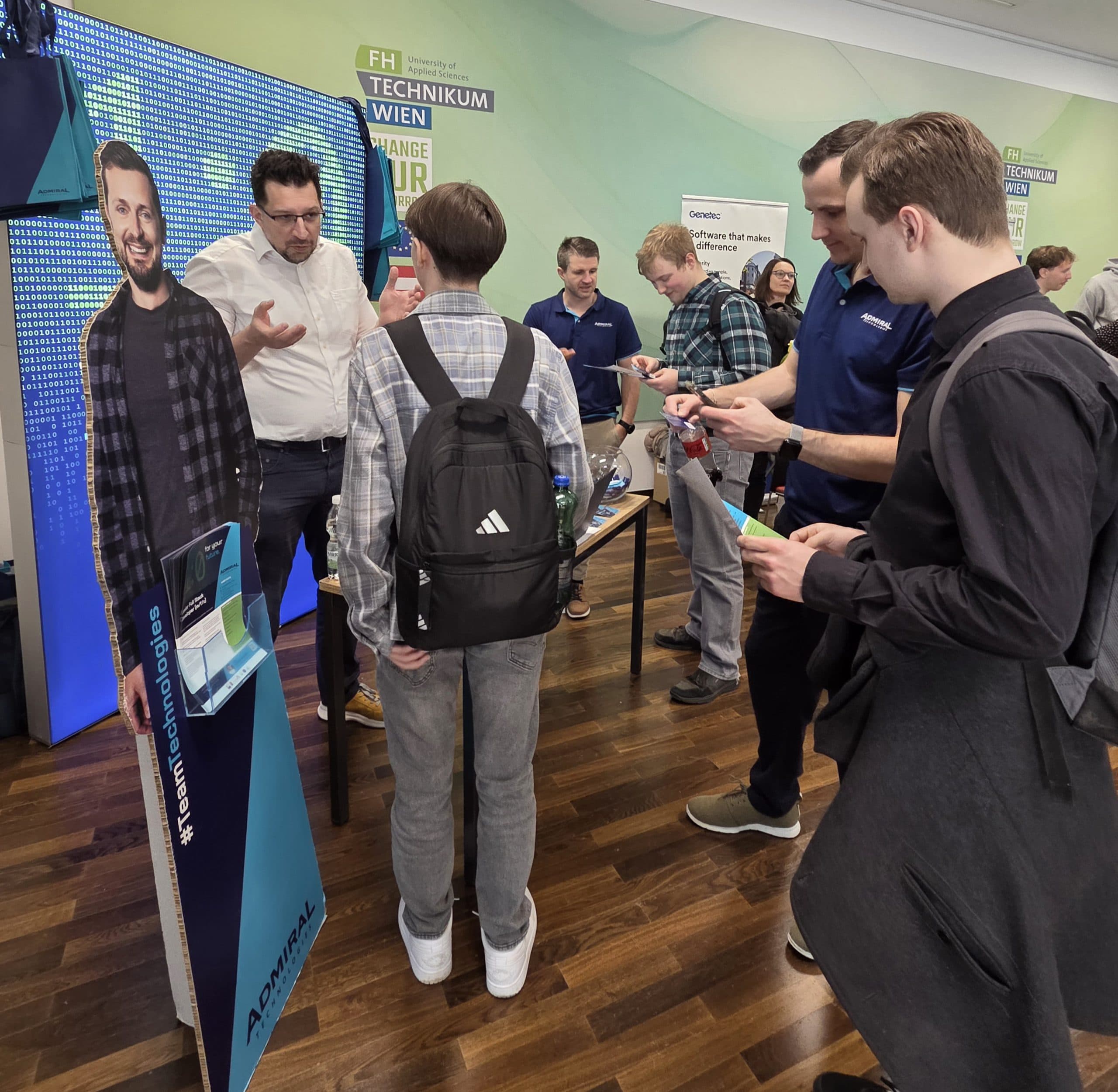 Mehrere Personen versammeln sich an einem Stand auf einer Technologiemesse und unterhalten sich mit den Ausstellern. Links steht ein Pappausschnitt eines lächelnden Mannes mit einem Laptop in der Hand. Der Boden ist aus Holz, und die Atmosphäre ist lebhaft.