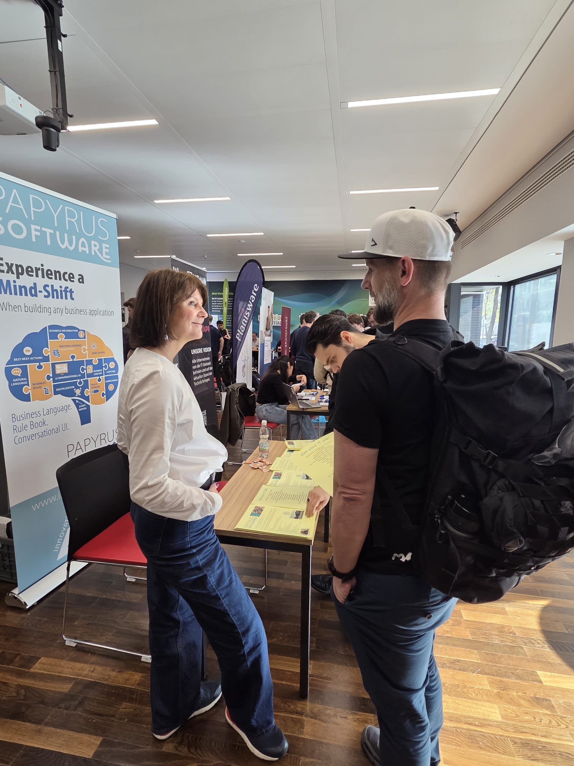 Eine Frau und ein Mann unterhalten sich am Stand eines Softwareunternehmens auf einer Jobmesse oder Expo. Andere Leute besuchen Stände im Hintergrund. Der Mann trägt einen großen schwarzen Rucksack. Der Stand zeigt ein Papyrus Software-Banner.