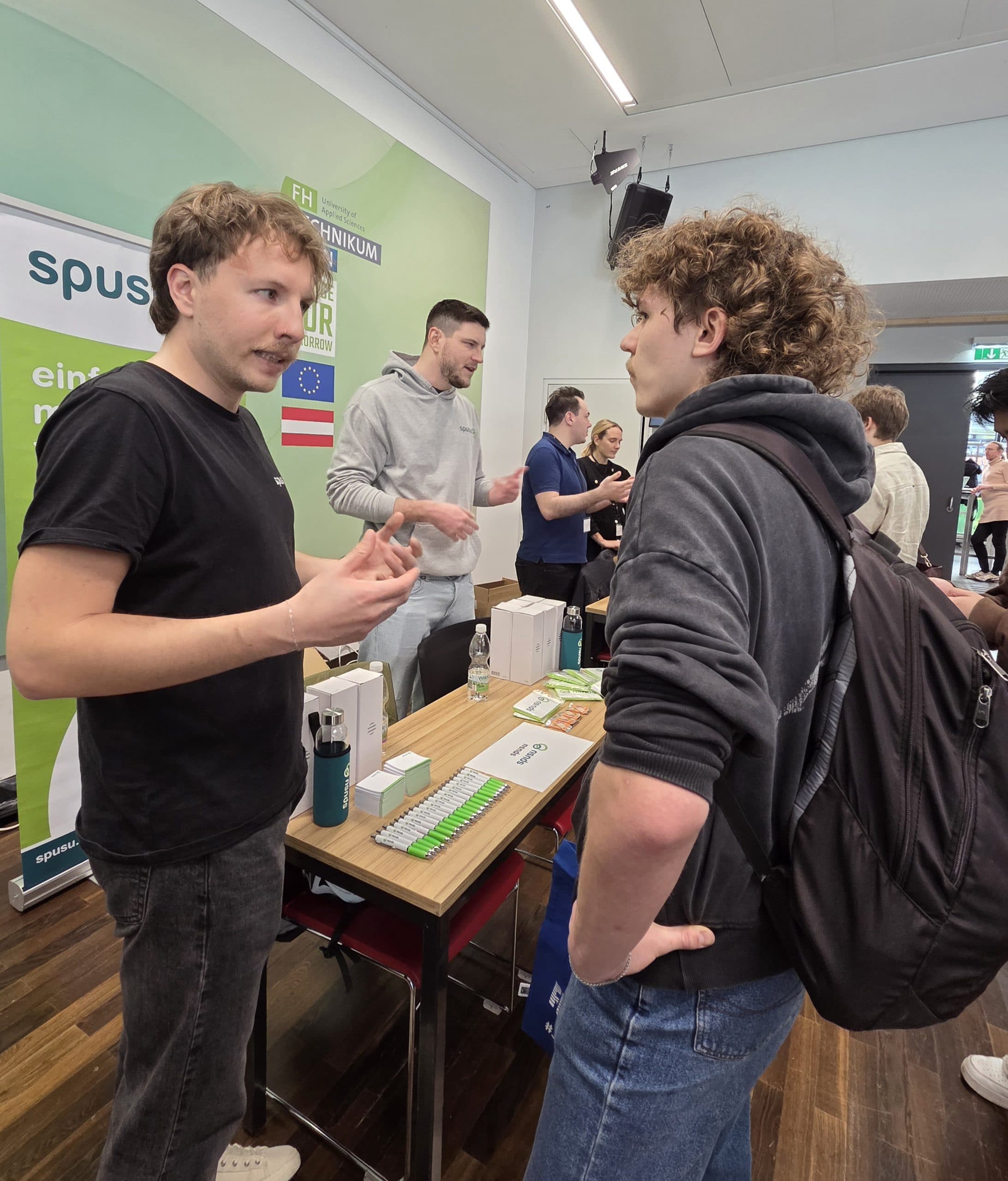 Zwei junge Männer unterhalten sich an einem Stand bei einer Indoor-Veranstaltung. Auf dem Tisch zwischen ihnen liegen Werbematerialien und kleine Schachteln. Im Hintergrund sind weitere Personen zu sehen, und hinter dem Stand sind Banner mit Text angebracht.