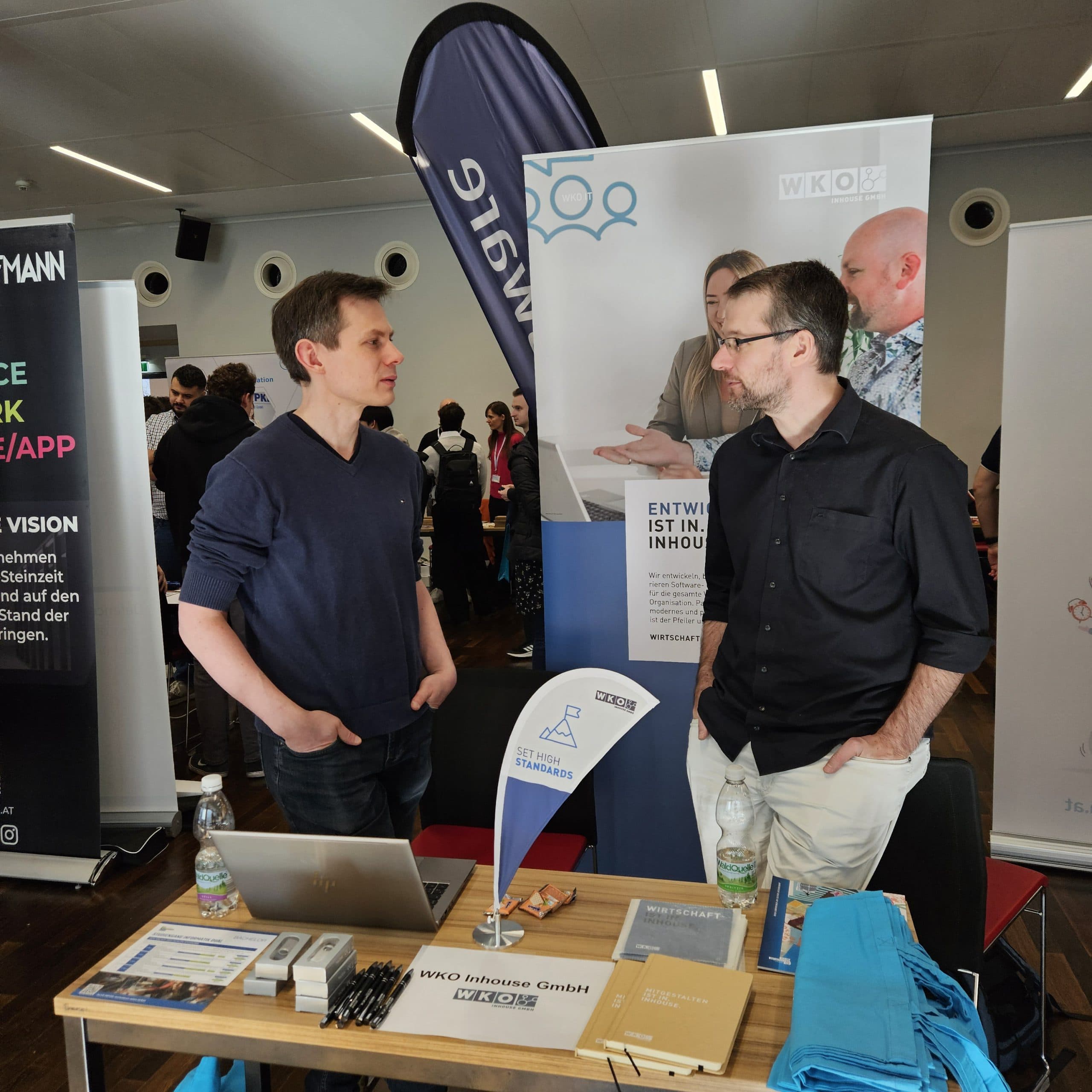 Zwei Männer stehen während einer Veranstaltung in einer Halle an einem Stand und unterhalten sich. Der Stand hat einen Laptop, Broschüren und Markenartikel auf dem Tisch. Hinter ihnen sind Banner und Informationsposter angebracht. Im Hintergrund sind andere Teilnehmer zu sehen.