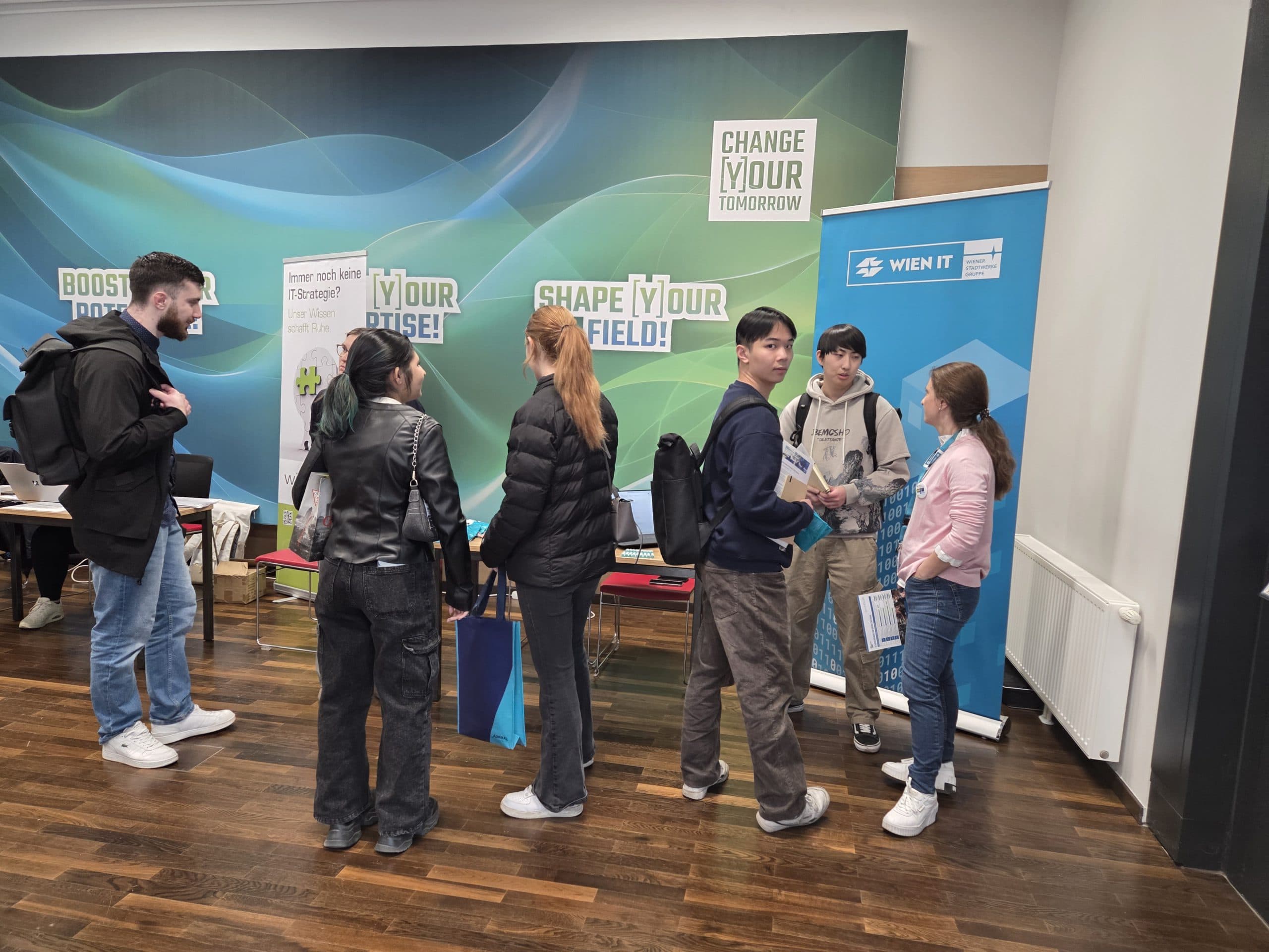 Eine Gruppe junger Leute mit Rucksäcken steht in der Schlange vor einem überdachten Stand mit Bannern, auf denen 'Boost your expertise', 'Shape your field' und 'Change your tomorrow' steht, an der Wand hinter ihnen.