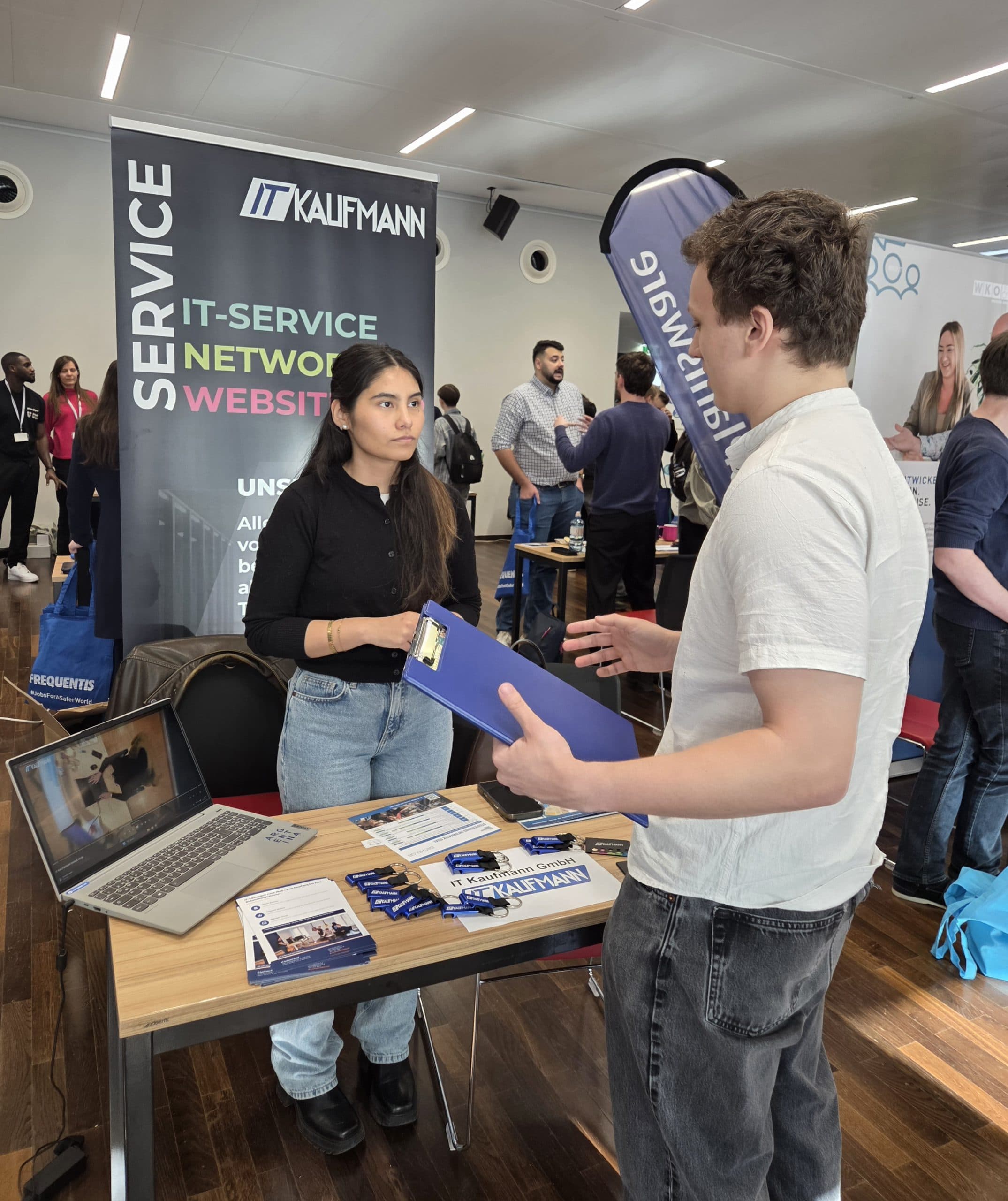Eine Frau steht hinter einem Standtisch mit Broschüren und Stiften und unterhält sich mit einem Mann, der ein Klemmbrett hält, auf einer Jobmesse. Hinter ihr ist ein Banner mit der Aufschrift 'IT Kalifmann Service IT-Service Network Website' zu sehen.