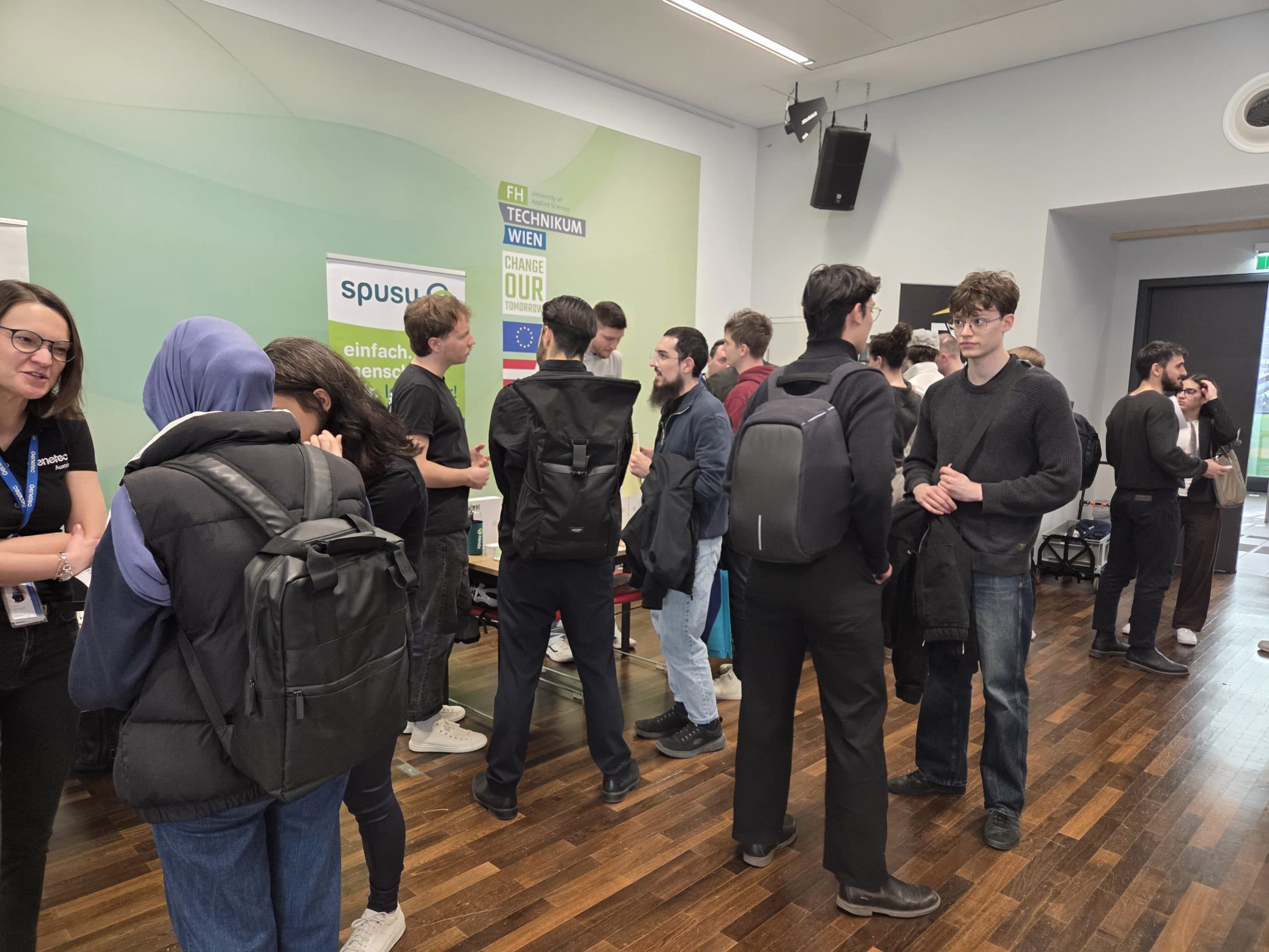Eine Gruppe von Studenten in legerer Kleidung steht in kleinen Gruppen bei einer Indoor-Veranstaltung mit Bannern im Hintergrund und unterhält sich. Der Raum ist mit Holzböden und hellgrünen Wänden ausgestattet.
