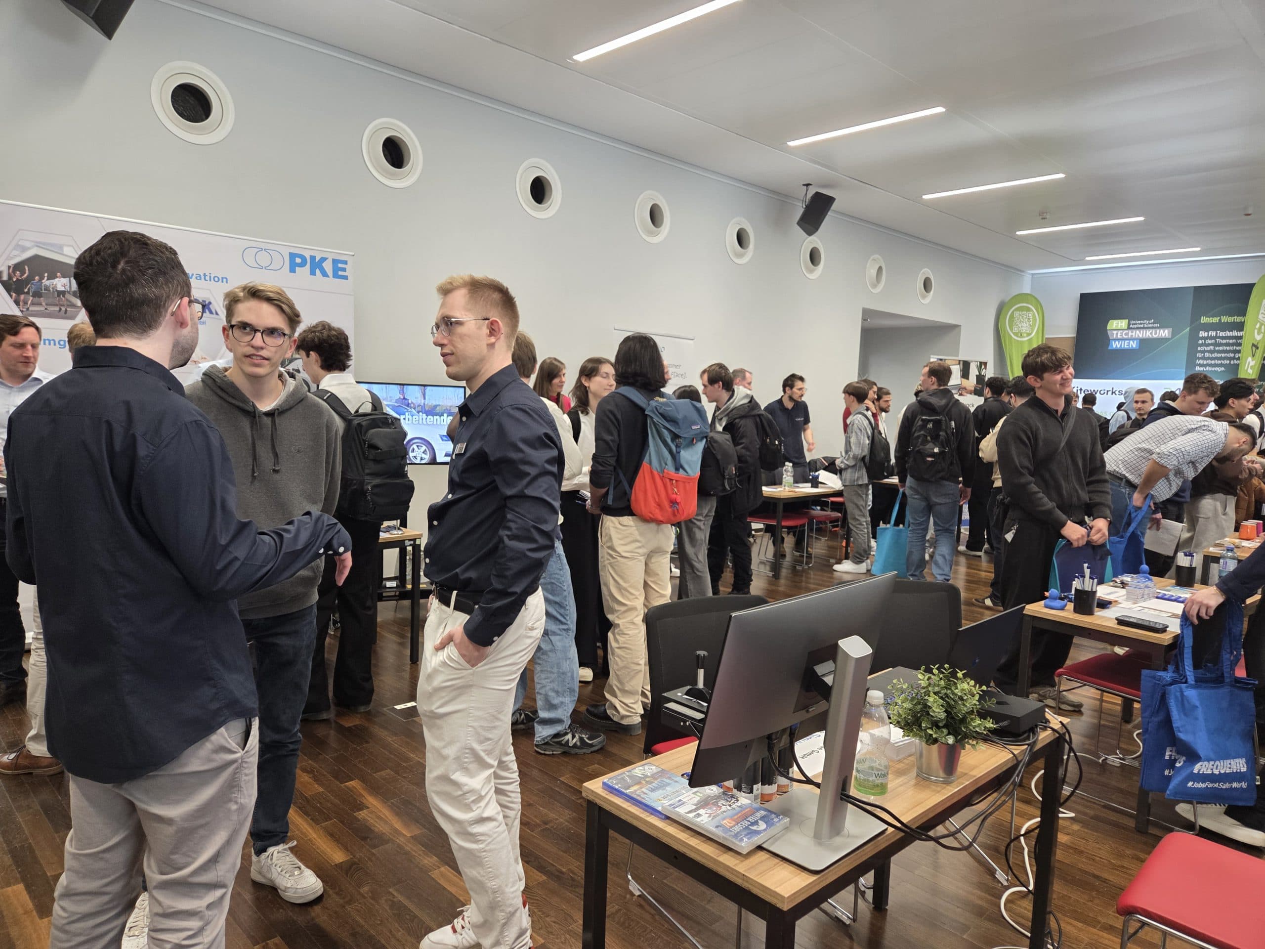 Eine Gruppe von Menschen interagiert auf einer belebten Tech-Messe oder einem Networking-Event, mit Informationsständen, Postern und Tischen mit Computern in einem modernen, gut beleuchteten Raum.