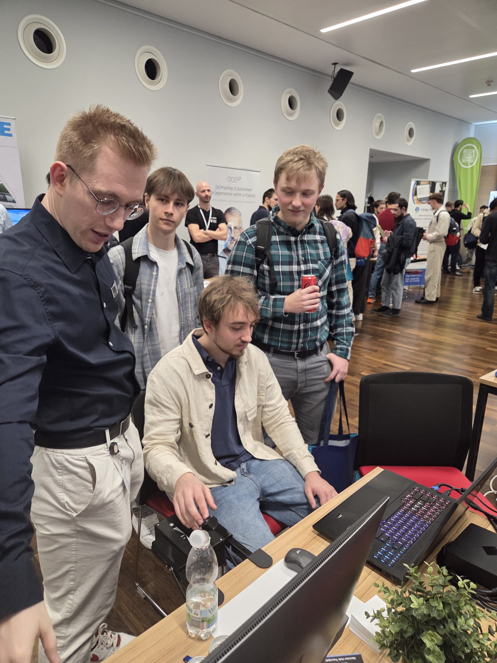 Vier junge Männer sind bei einer Veranstaltung oder Konferenz in einem Gebäude um einen Computer versammelt. Einer sitzt und benutzt die Maus, während die anderen stehen und zusehen. Im Hintergrund sind Menschen und Messestände zu sehen.