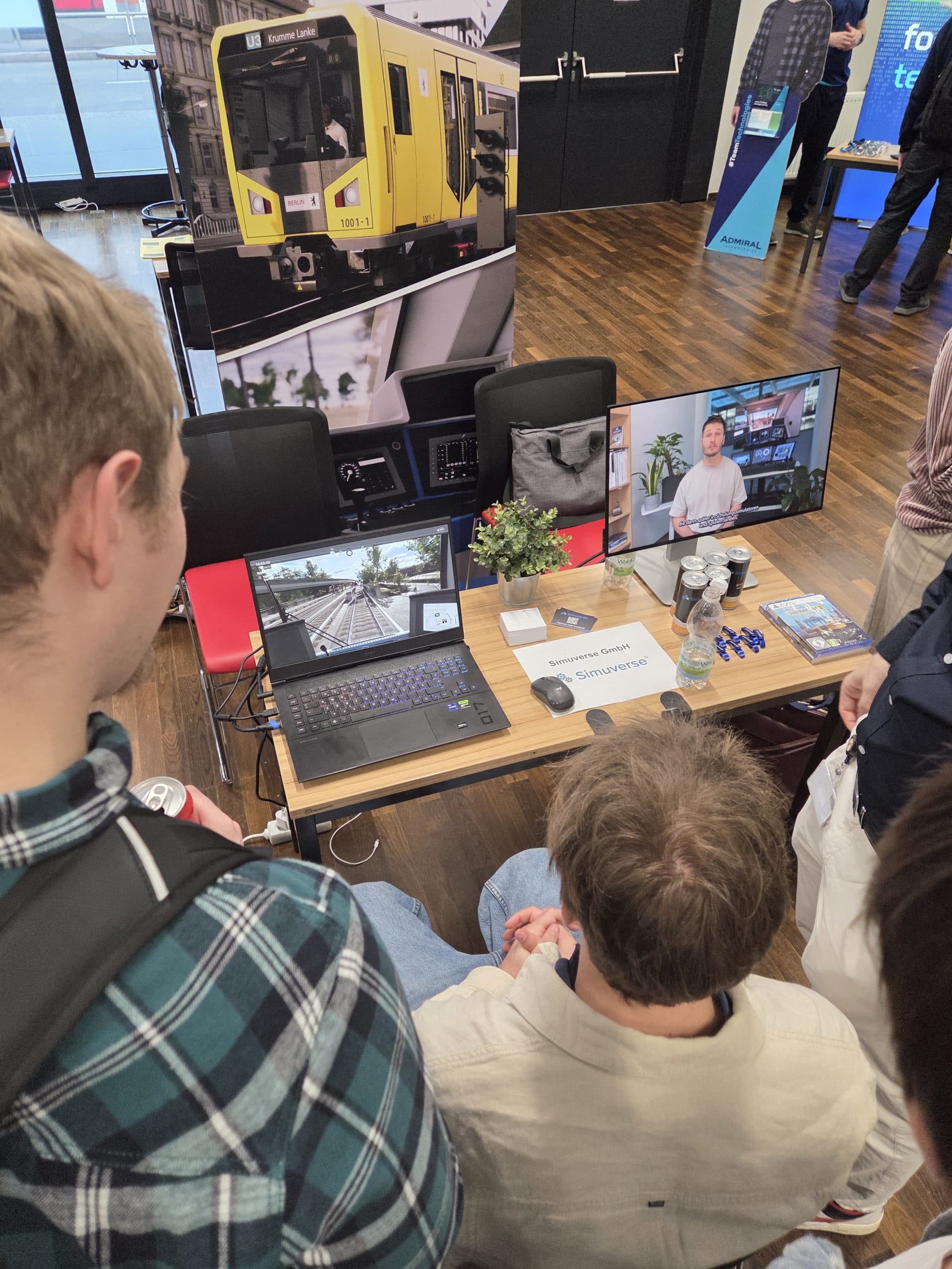 Zwei Personen schauen auf einen Tisch mit einem Laptop, einem Monitor und einer Zugfahrsimulationsanlage bei einer Indoor-Veranstaltung. Auf den Bildschirmen sind Zugsteuerungen und ein Sprecher auf Video zu sehen. Auf dem Tisch befinden sich Werbeartikel und Broschüren.