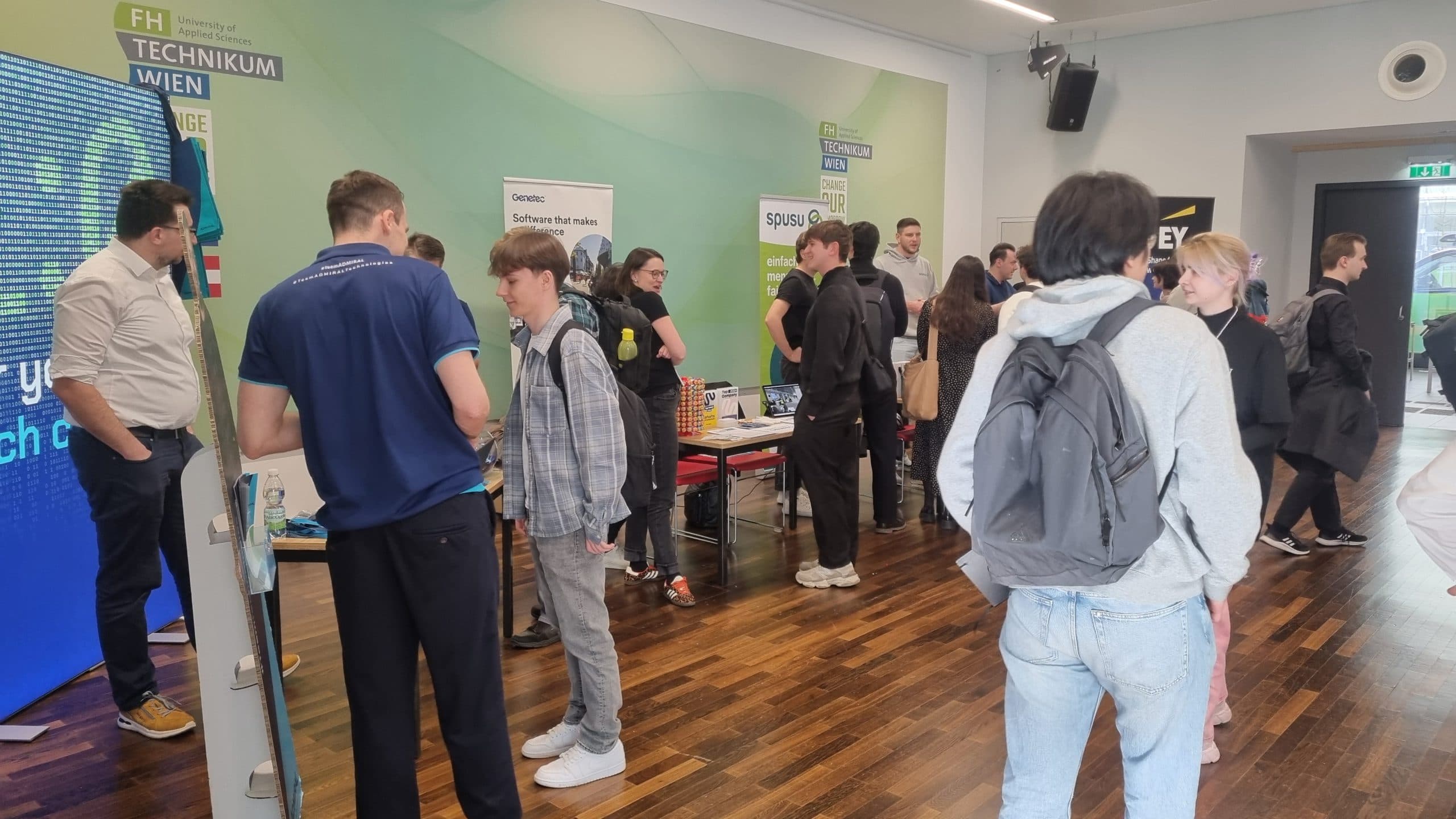 Eine Gruppe von Menschen interagiert auf einer technischen Veranstaltung in einer Halle mit mehreren Ständen, Postern und Informationsständen. Die Teilnehmer sind in Gespräche vertieft und einige begutachten die Auslagen. Die Atmosphäre ist lebhaft und professionell.