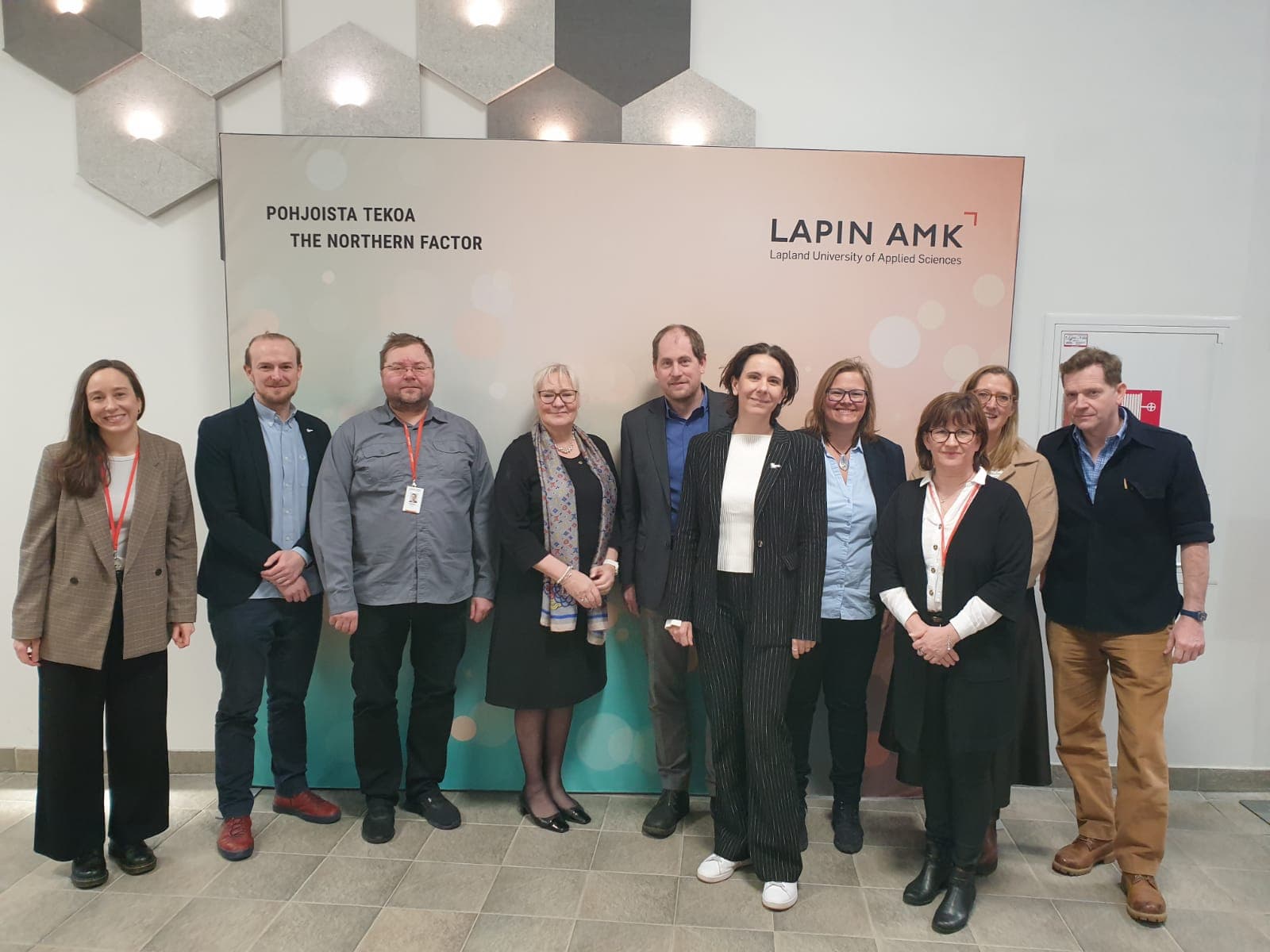 Eine Gruppe von zehn Personen steht in einem Innenraum vor einem Banner der Lapland University of Applied Sciences und lächelt in die Kamera. Der Hintergrund besteht aus geometrischen Wandtafeln und Text in Finnisch und Englisch.