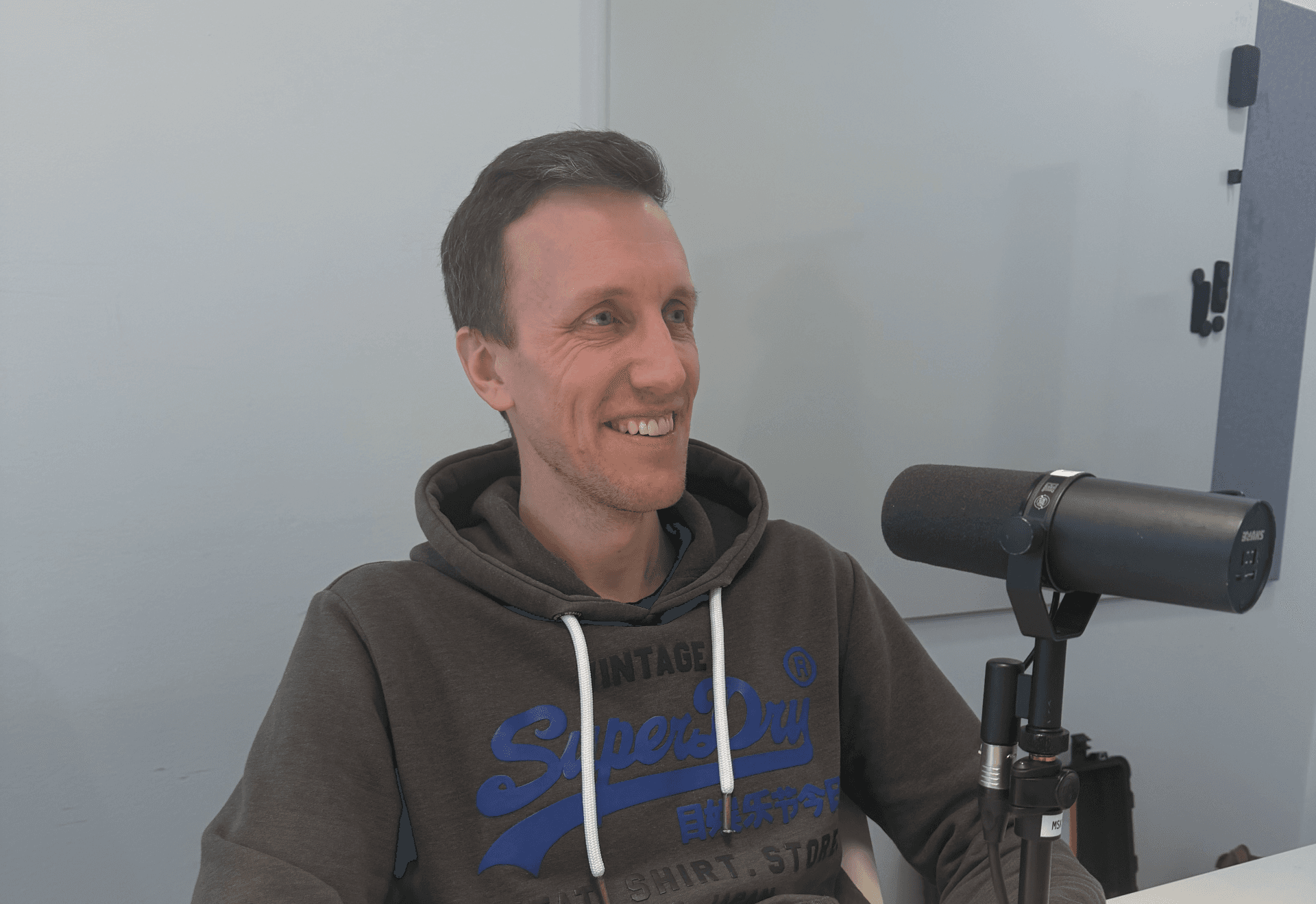Ein lächelnder Mann in einem dunklen Kapuzenpulli sitzt an einem Tisch mit einem Mikrofon auf einem Ständer vor ihm und scheint einen Podcast oder ein Interview aufzunehmen oder zu sprechen.
