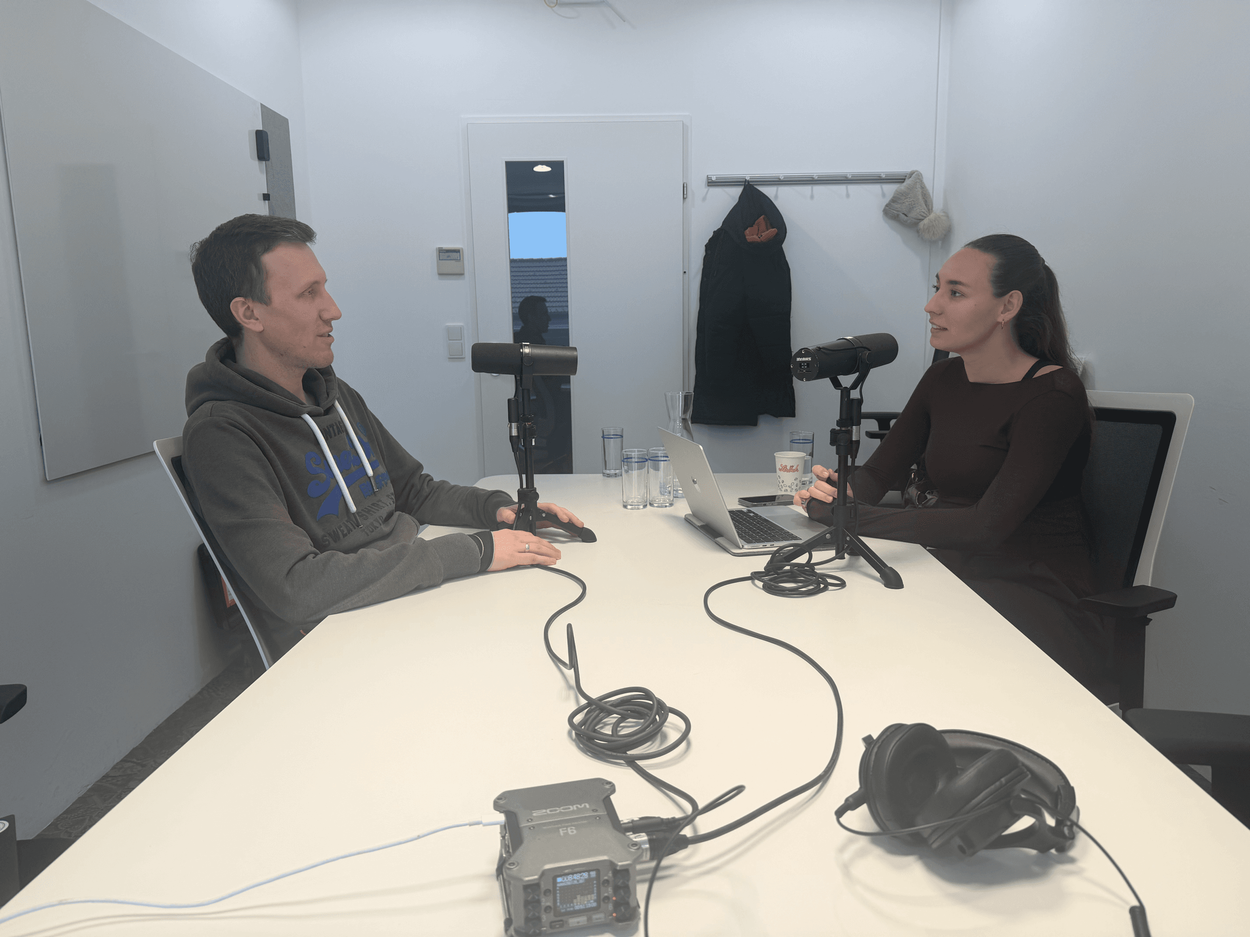 Zwei Personen sitzen sich an einem Tisch in einem modernen Büro gegenüber und sprechen in Mikrofone. Auf dem Tisch stehen ein Laptop, Wassergläser, Kopfhörer und Audiogeräte. An der Wand hinter ihnen hängen Mäntel.
