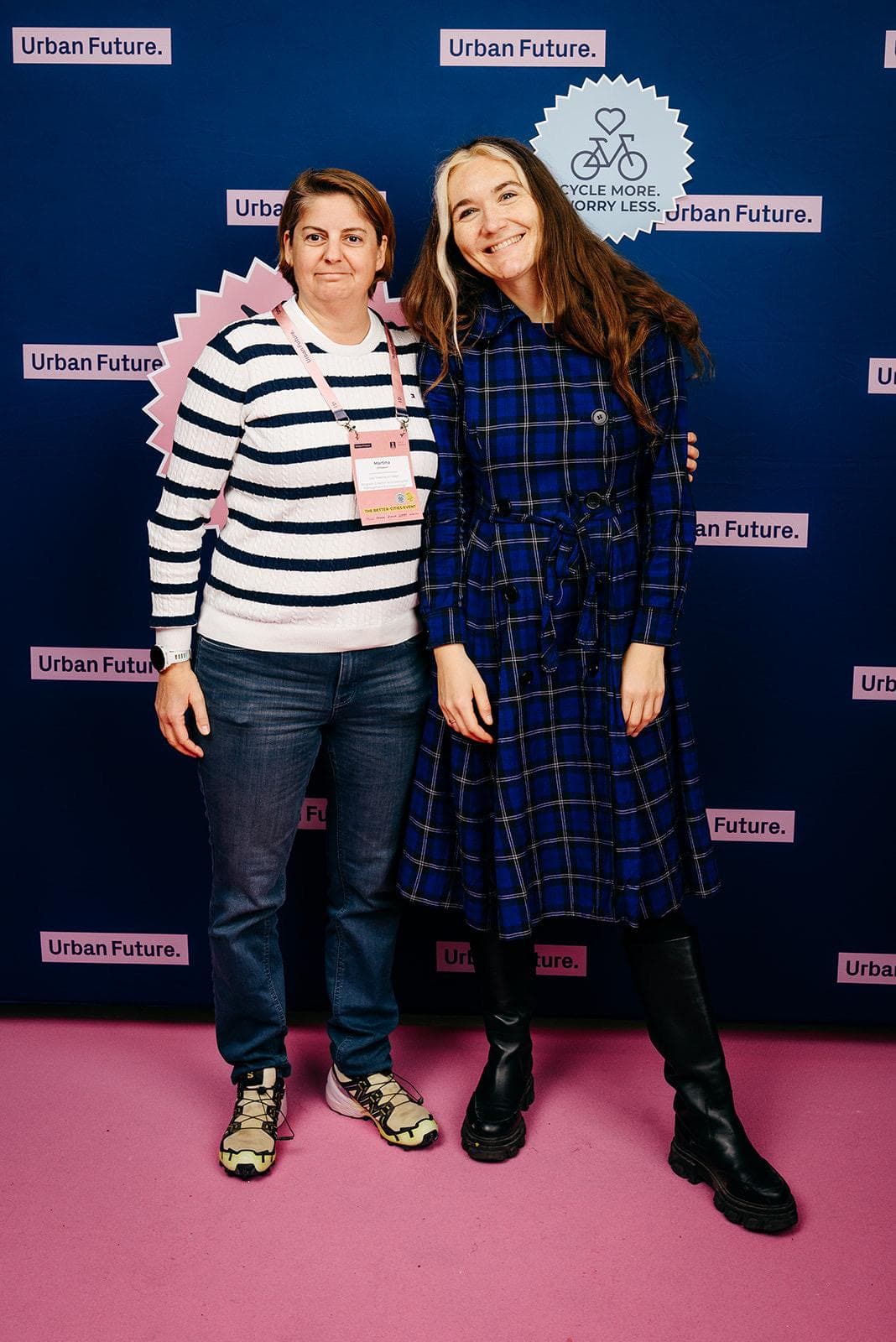 Zwei Personen stehen lächelnd vor einem blauen Hintergrund mit Urban Future-Logos. Die eine trägt einen gestreiften Pullover und Jeans, die andere ein blau kariertes Kleid und Stiefel. Beide posieren auf einem rosa Boden.