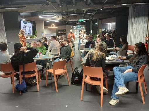 Eine Gruppe von Menschen sitzt an Tischen in einem modernen Innenraum und hört einer Frau zu, die an der Stirnseite neben einer projizierten Leinwand spricht. Einige Leute essen gerade, und die Atmosphäre wirkt locker und engagiert.