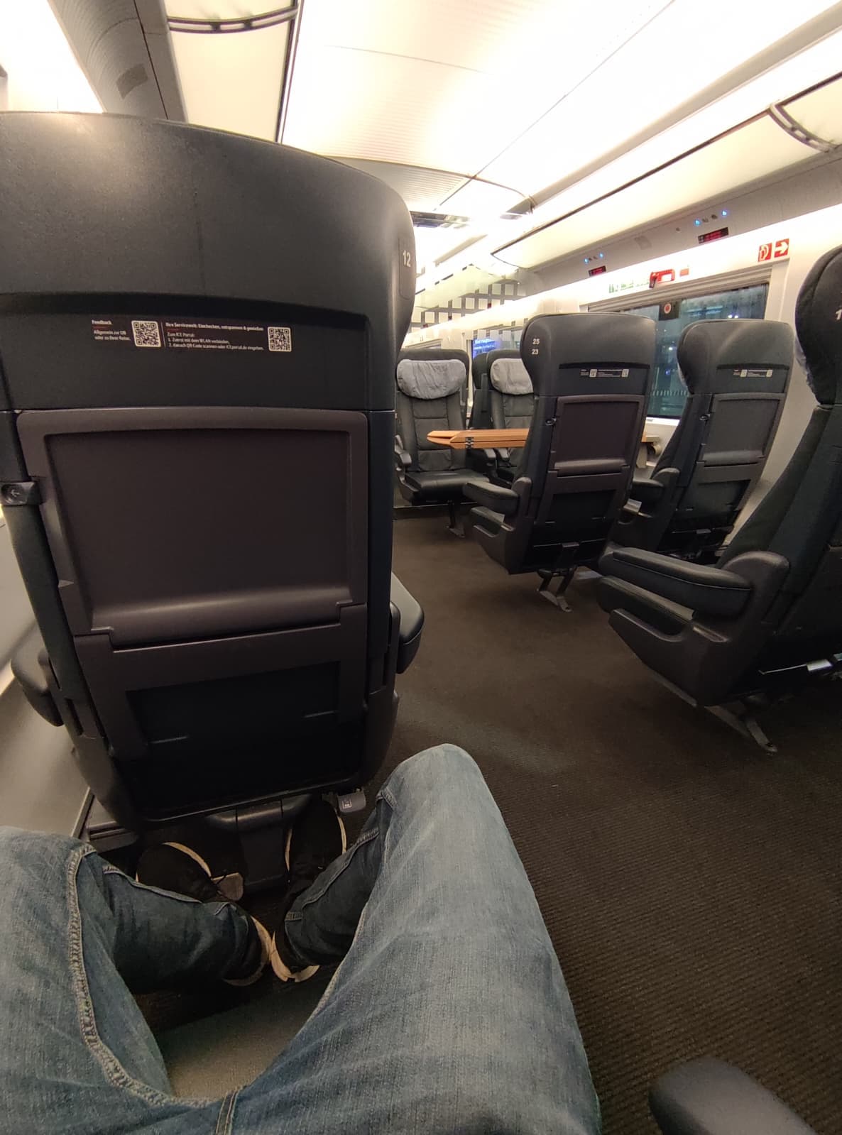 Eine Person in Jeans und Turnschuhen sitzt in einem modernen Zug mit großen, leeren schwarzen Sitzen und einem kleinen Tisch in der Mitte. Die Ansicht ist aus der Perspektive der Person und zeigt ihre ausgestreckten Beine.